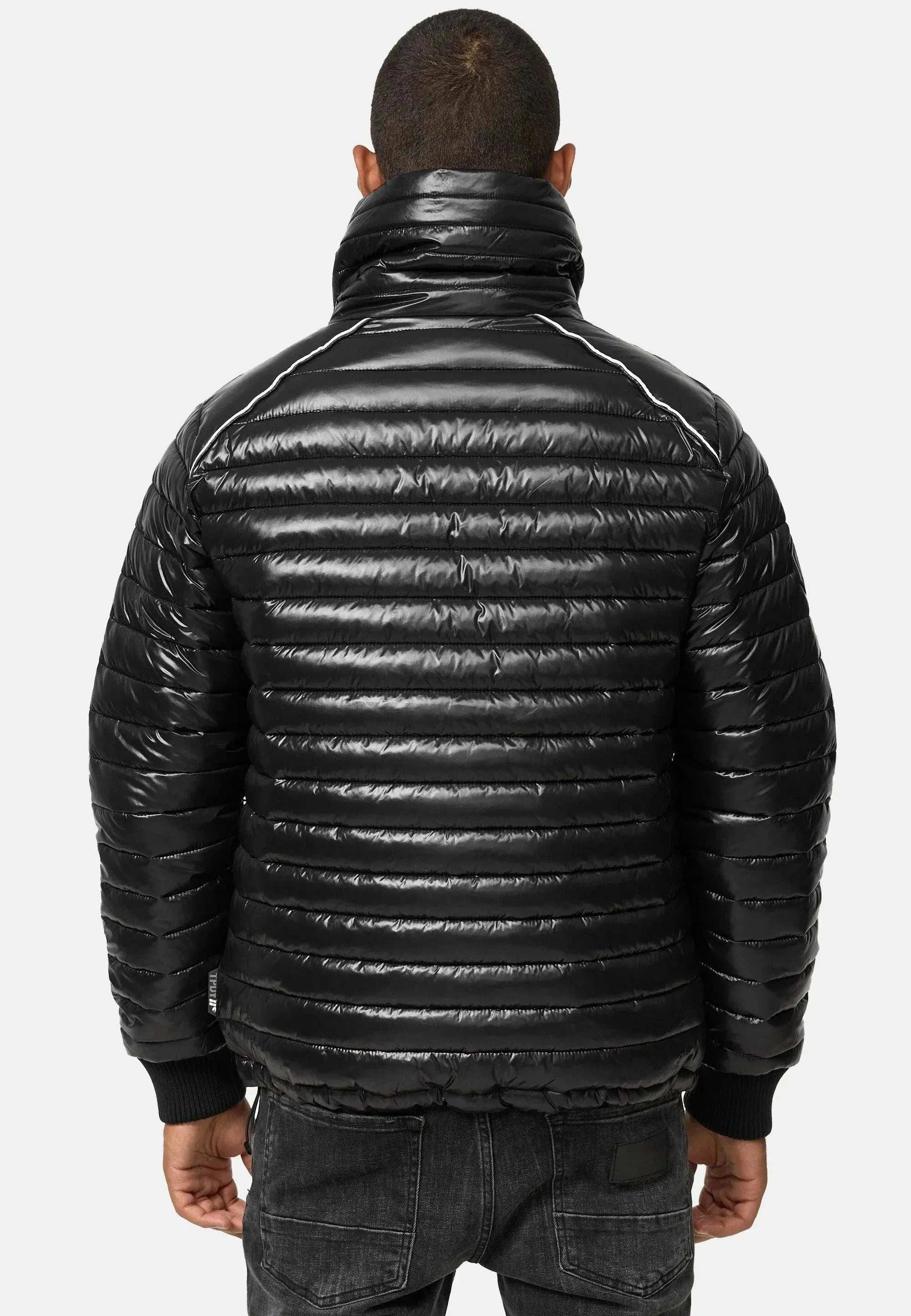 Eine Person ist von hinten zu sehen. Sie trägt eine glänzende Trueprodigy Corey Steppjacke mit Stehkragen mit horizontaler Steppung. Zusammen mit dunklen Jeans bildet die schlanke Silhouette einen Kontrast zum schlichten weißen Hintergrund.