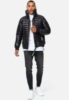 Ein Mann in einer Steppjacke mit Stehkragen von trueprodigy Corey, weißem T-Shirt, dunklen Jeans und weißen Turnschuhen schreitet vorwärts. Er hält mit der linken Hand den Reißverschluss der Jacke fest und steht vor einem schlichten hellgrauen Hintergrund.