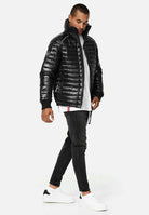 Die trueprodigy Corey Steppjacke mit Stehkragen lässt sich mühelos mit einem weißen T-Shirt, dunklen Röhrenjeans und weißen Sneakers kombinieren. Sie stehen vor einem hellgrauen Hintergrund, leicht angewinkelt, mit den Händen an den Seiten.