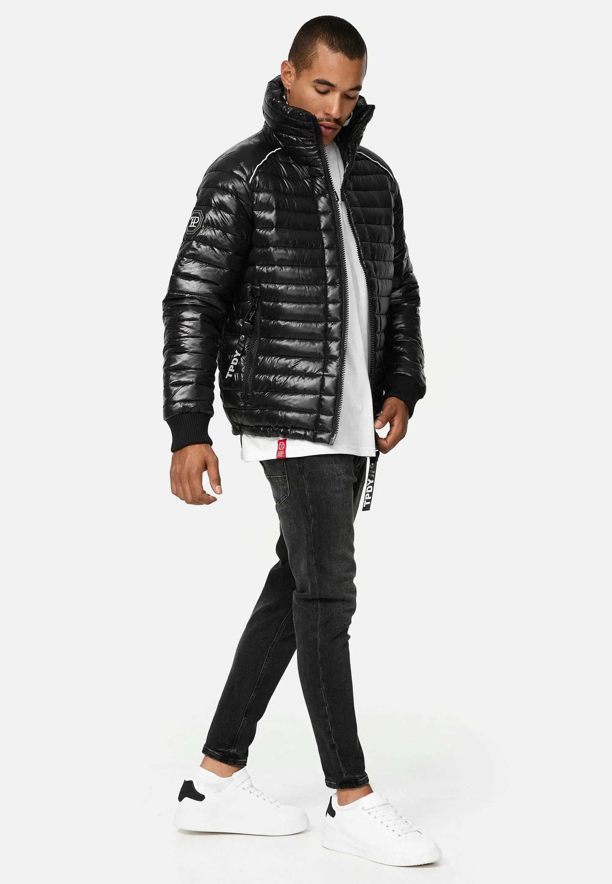 Die trueprodigy Corey Steppjacke mit Stehkragen lässt sich mühelos mit einem weißen T-Shirt, dunklen Röhrenjeans und weißen Sneakers kombinieren. Sie stehen vor einem hellgrauen Hintergrund, leicht angewinkelt, mit den Händen an den Seiten.