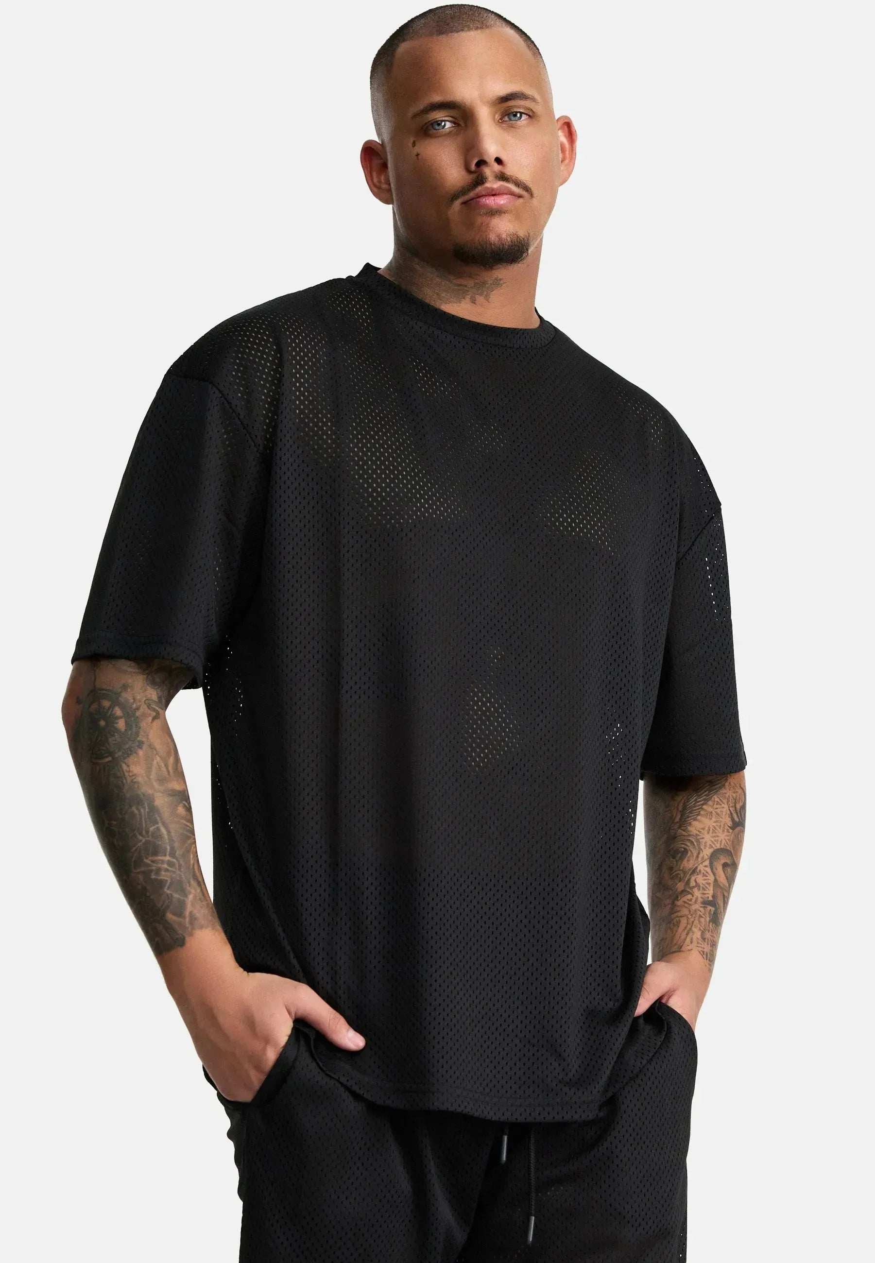 Der tätowierte Mann verströmt Vintage-Vibes vor einem schlichten Hintergrund. Er trägt das Daniel Oversized T-Shirt mit Druck und die dazu passende Hose von trueprodigy. Die Hände in den Taschen, behält er einen neutralen Gesichtsausdruck.