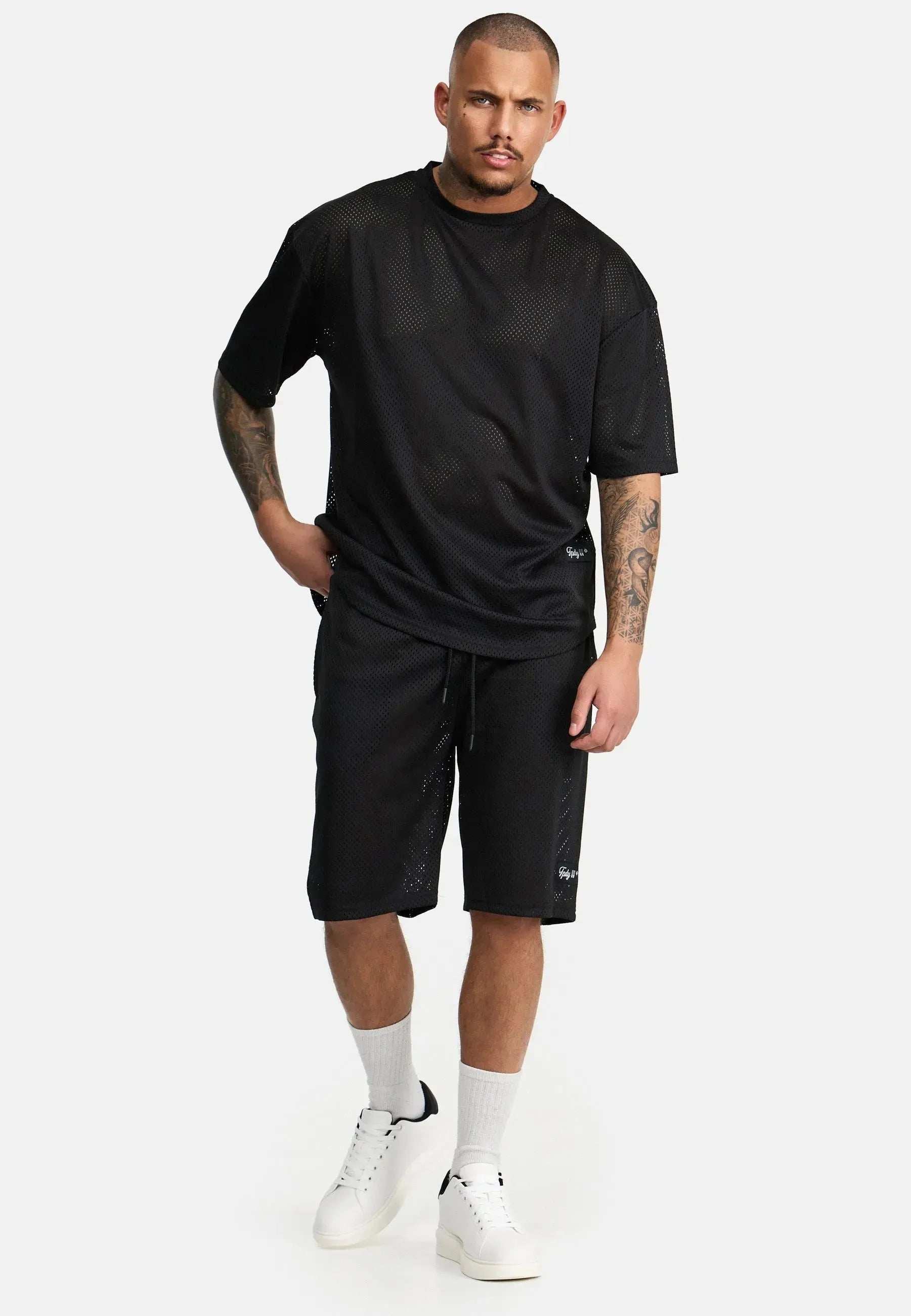 Vor einem schlichten Hintergrund steht ein Mann und trägt ein schwarzes Daniel Oversized T-Shirt mit Druck von trueprodigy, dazu passende Shorts. Mit einer Hand in der Tasche vervollständigt er seinen Vintage-Look mit weißen Sneakers und Socken.