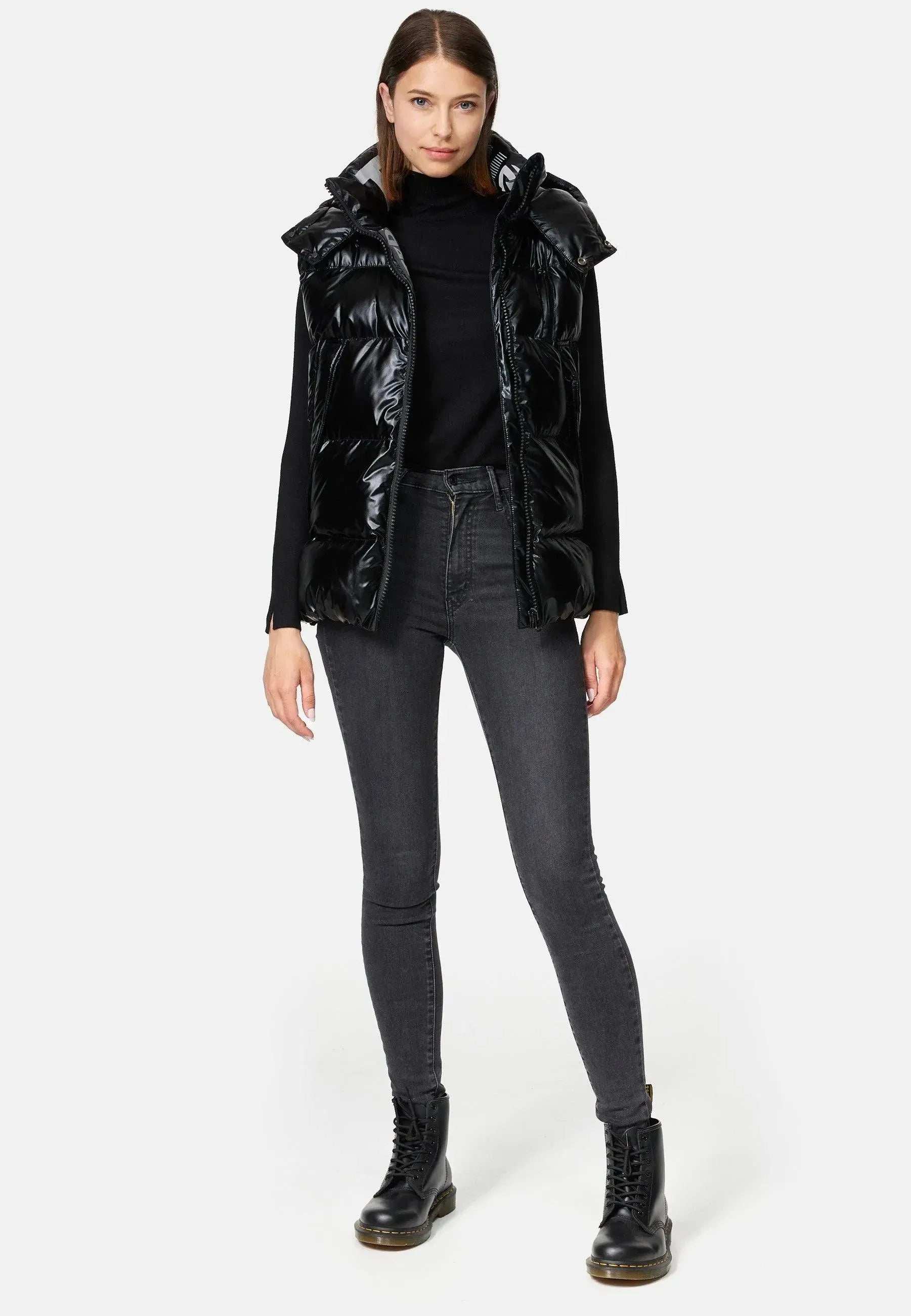 Eine Frau steht vor einem schlichten Hintergrund und trägt eine Trueprodigy Darko F Weste glänzend mit Kapuze über einem schwarzen Rollkragenpullover, dazu dunkelgraue Röhrenjeans und schwarze Stiefel. Ihre Hände ruhen an ihren Seiten und zeigen einen neutralen Gesichtsausdruck.