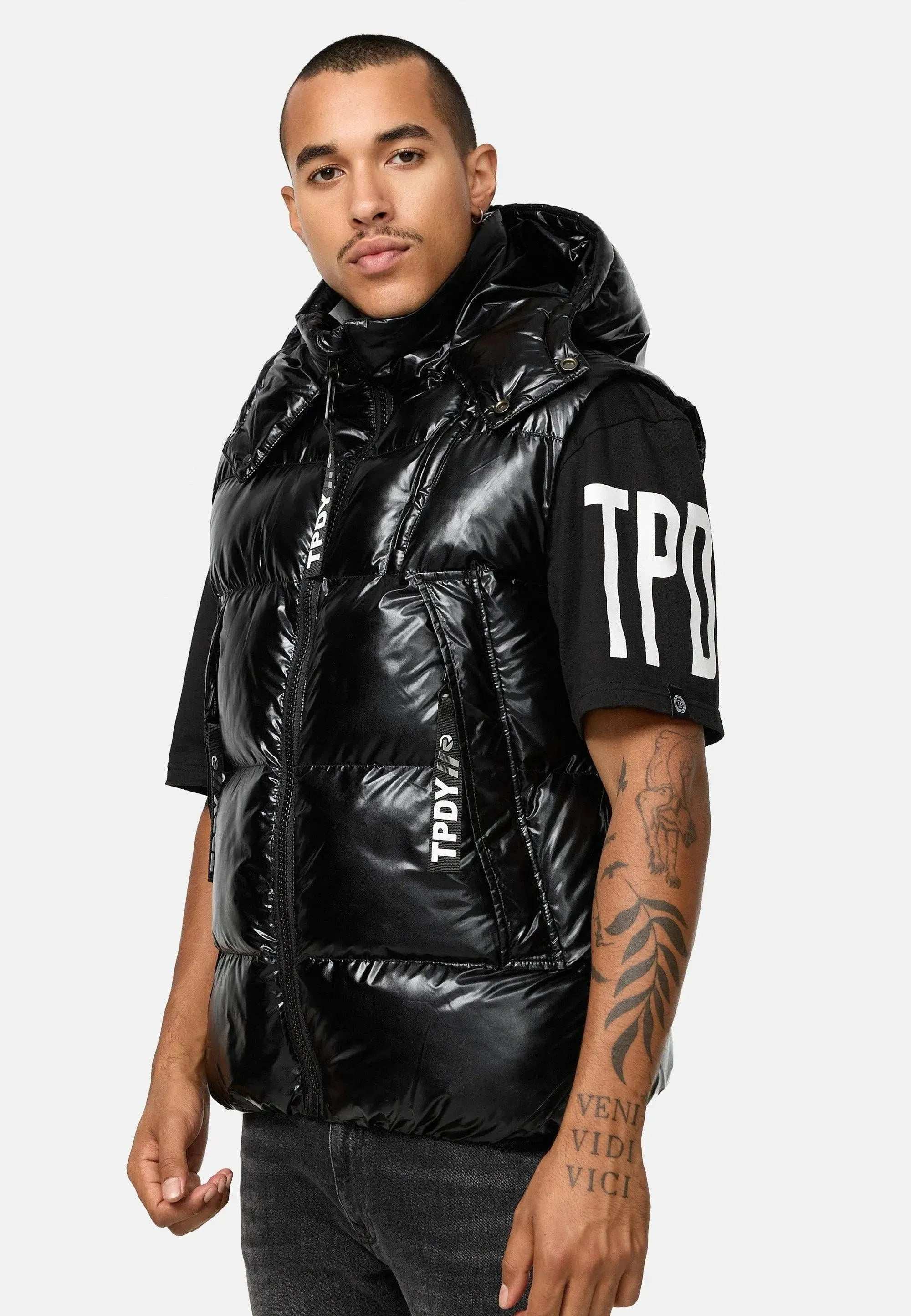 Eine Person mit kurzen Haaren trägt eine Trueprodigy Darko Steppweste glänzend mit Kapuze über einem Trueprodigy TPD T-Shirt. Die Weste mit abnehmbarer Kapuze ist mit einem Unterarmtattoo vor einem schlichten weißen Hintergrund kombiniert.