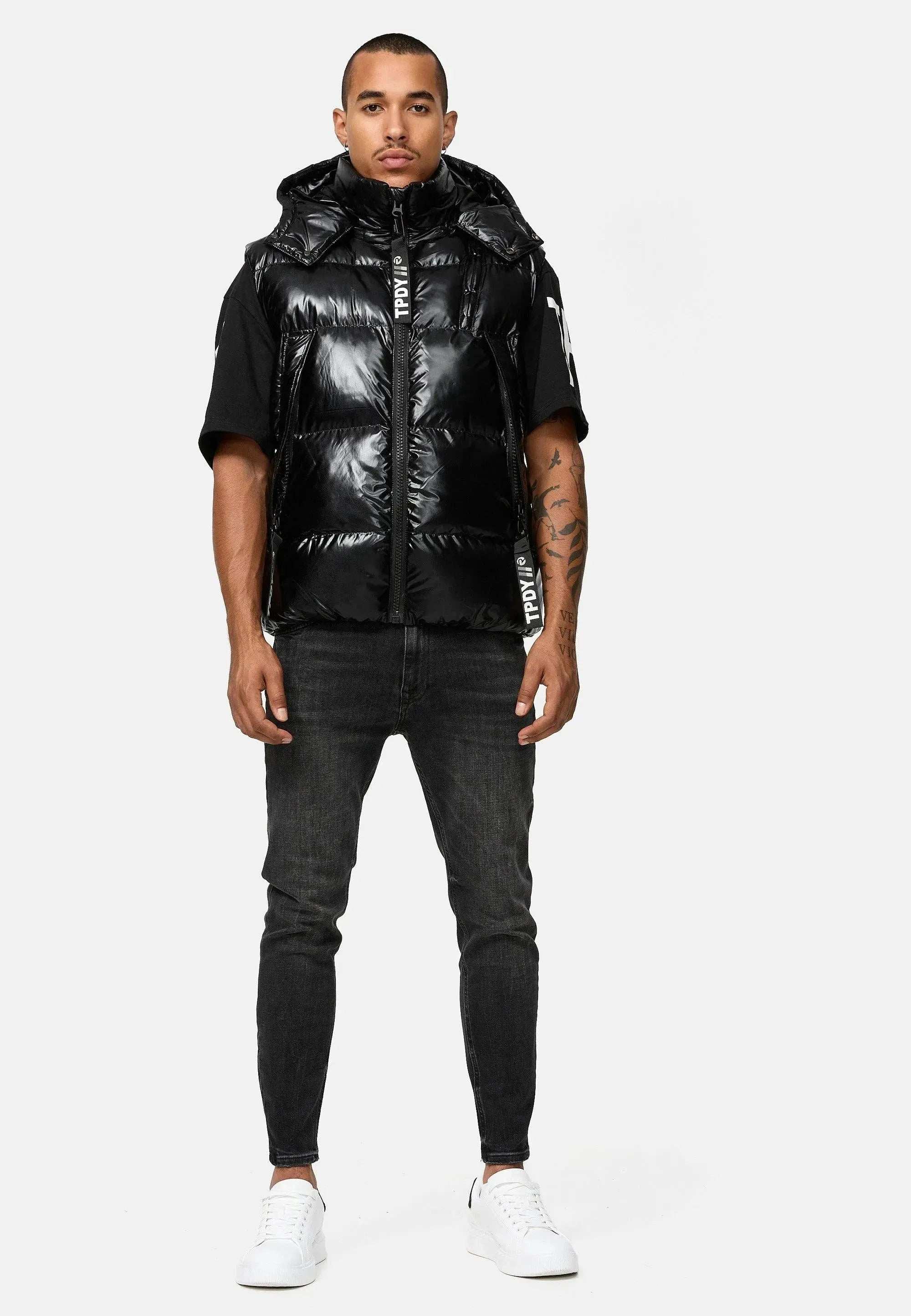 Vor einem schlichten Hintergrund trägt eine Person trueprodigys glänzend schwarze Darko Steppweste mit Kapuze über einem schwarzen Kurzarmhemd, dunklen Jeans und weißen Turnschuhen. Tattoos zieren ihre Arme, während sie einen neutralen Gesichtsausdruck wahren.