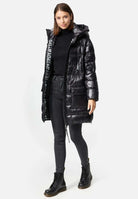 Eine Frau steht vor einem schlichten Hintergrund in einer glänzenden David F Glänzende Steppjacke mit Kapuze von trueprodigy, elegant kombiniert mit einem schwarzen Rollkragenpullover, dunklen Röhrenjeans und eleganten schwarzen Stiefeln. Ihr leichtes Lächeln und die rechte Hand, die lässig in ihrer Manteltasche steckt, strahlen Selbstvertrauen und Stil aus.