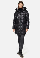 Eine Person trägt einen langen schwarzen Damenparka mit Text auf den Reißverschlüssen, über einem schwarzen Rollkragenpullover und dunklen Jeans. Der Look wird durch stylische schwarze Schnürstiefel komplettiert. Sie stehen vor einem schlichten weißen Hintergrund. (Produkt: David F Glänzende Steppjacke mit Kapuze von trueprodigy).