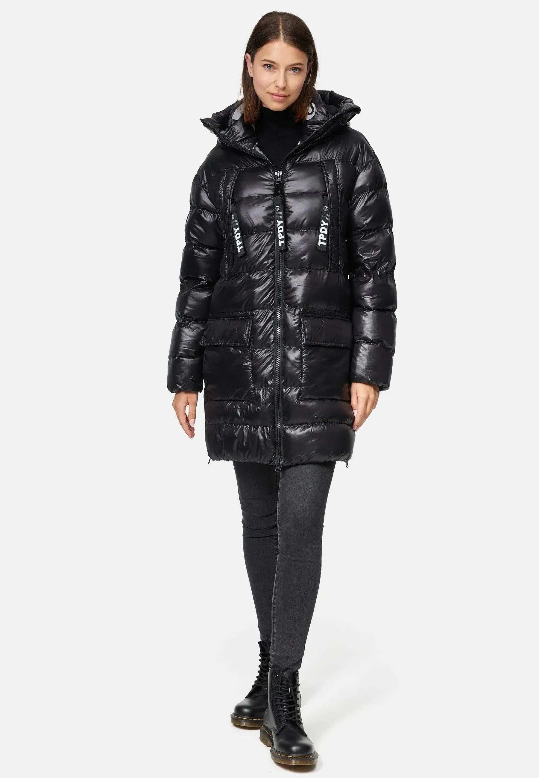 Eine Person trägt einen langen schwarzen Damenparka mit Text auf den Reißverschlüssen, über einem schwarzen Rollkragenpullover und dunklen Jeans. Der Look wird durch stylische schwarze Schnürstiefel komplettiert. Sie stehen vor einem schlichten weißen Hintergrund. (Produkt: David F Glänzende Steppjacke mit Kapuze von trueprodigy).