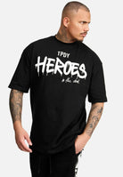 Ein tätowierter Mann trägt ein übergroßes schwarzes Dexter-T-Shirt von trueprodigy aus schwerem Jersey. Darunter ist fettgedruckt „in the dark“ zu sehen. Er blickt nach links, seine rechte Hand steckt lässig in der Tasche seiner schwarzen Hose.