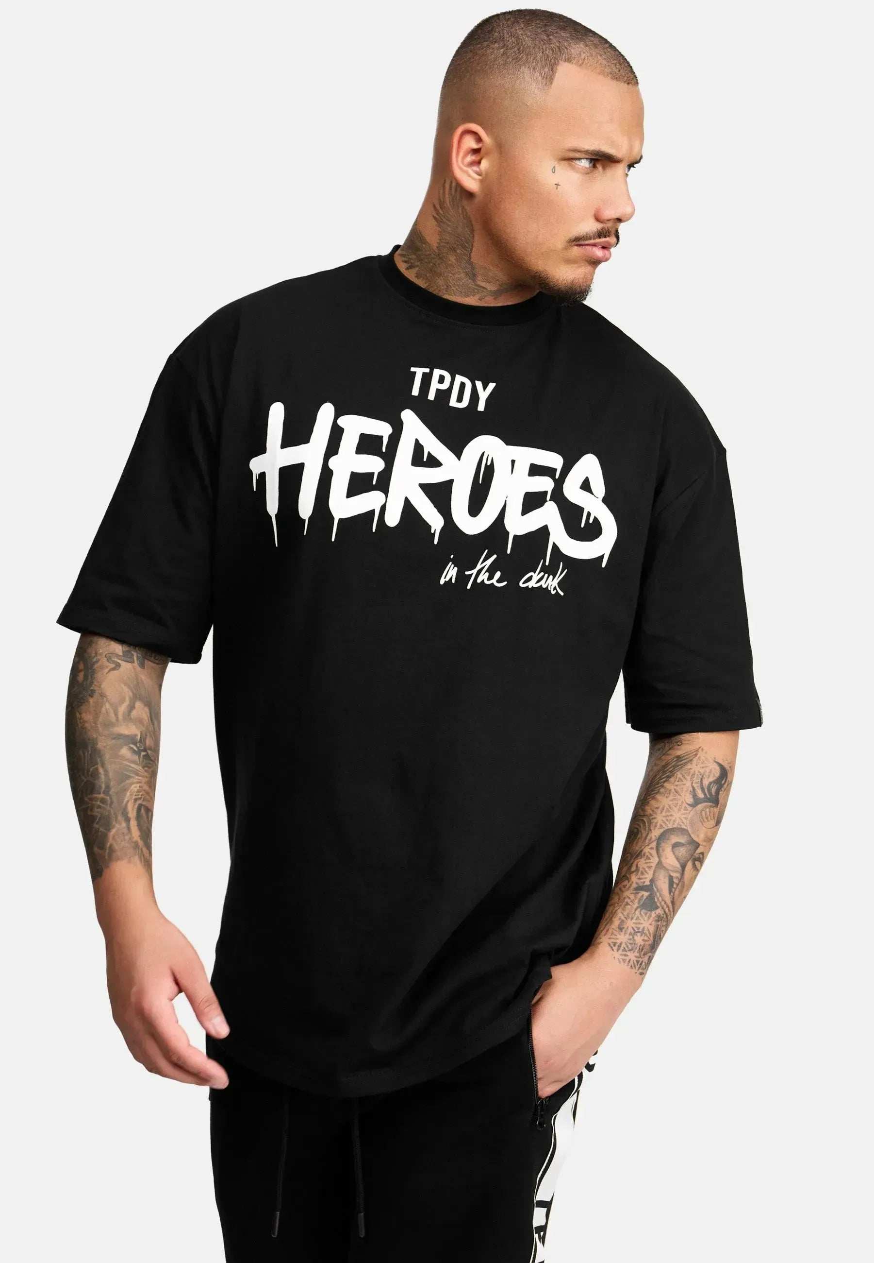 Ein tätowierter Mann trägt ein übergroßes schwarzes Dexter-T-Shirt von trueprodigy aus schwerem Jersey. Darunter ist fettgedruckt „in the dark“ zu sehen. Er blickt nach links, seine rechte Hand steckt lässig in der Tasche seiner schwarzen Hose.