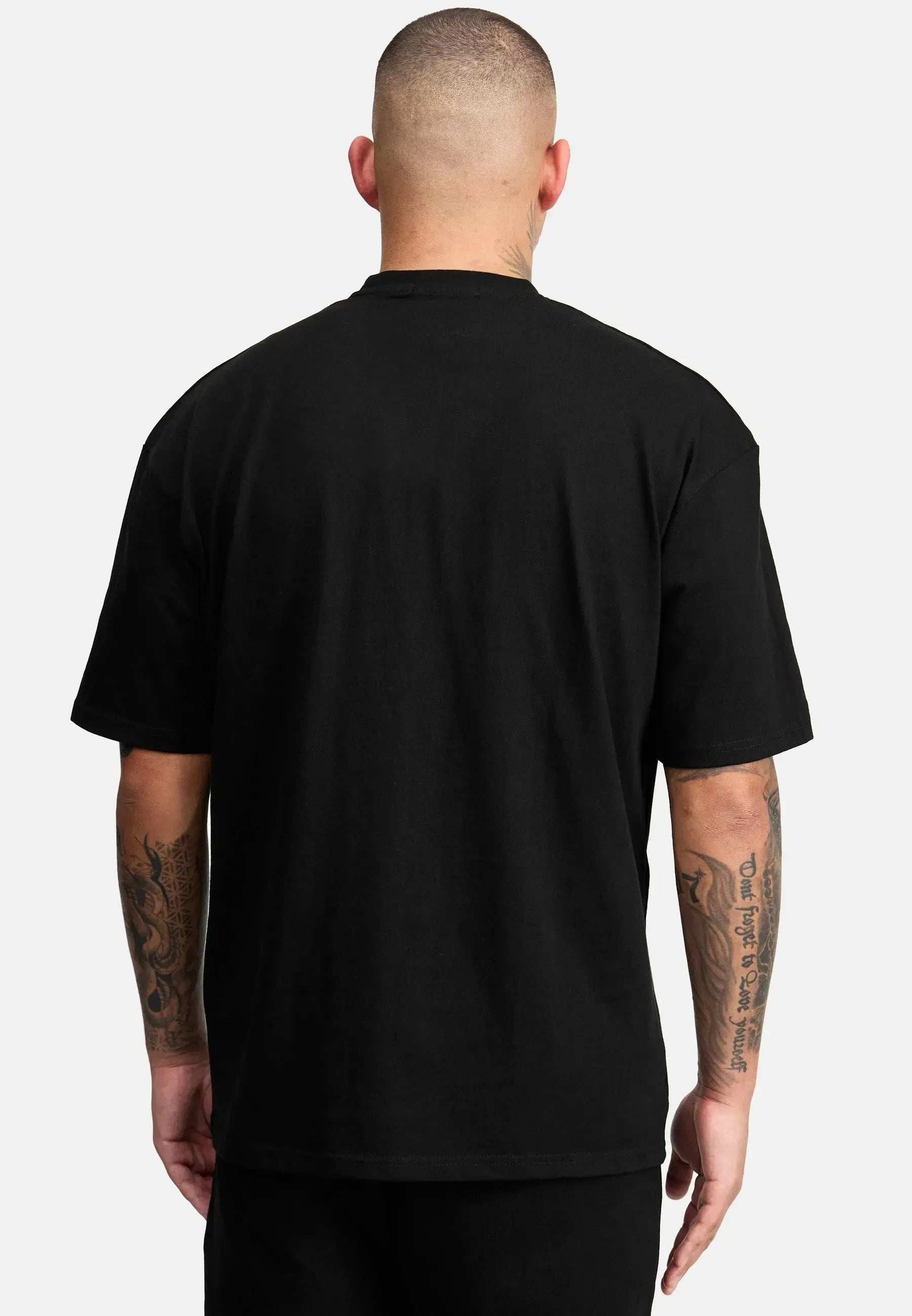Ein Mann mit rasiertem Kopf steht mit dem Rücken zur Kamera. Er trägt ein Dexter Oversized T-Shirt mit Druck von trueprodigy und eine schwarze Hose vor einem schlichten weißen Hintergrund. Auf beiden Armen sind Tattoos zu sehen.