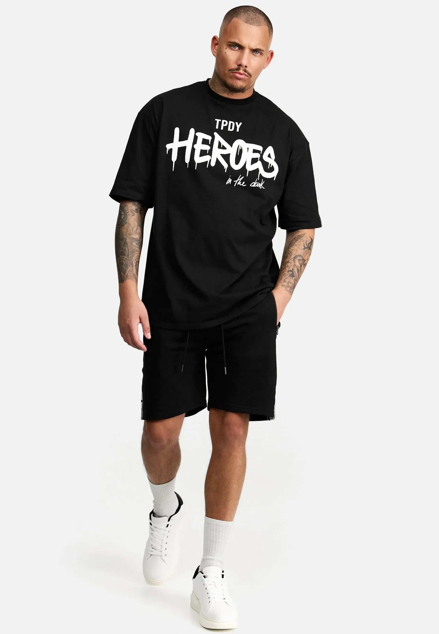 Ein tätowierter Mann schreitet in einem übergroßen schwarzen Dexter-T-Shirt von trueprodigy mit dem weißen Text „Heroes in the dark“ vorwärts. Er kombiniert es mit schwarzen Shorts, weißen Socken und Turnschuhen. Der schlichte hellgraue Hintergrund unterstreicht seinen kühnen Look.