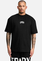 Eine tätowierte Person trägt ein übergroßes schwarzes T-Shirt mit „TPDY“ aus der Elijah-Linie von Trueprodigy, dazu eine schwarze Hose auf weißem Hintergrund.