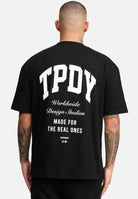 Eine Person mit rasiertem Kopf und Tattoos auf dem linken Arm trägt ein schwarzes „Elijah Oversized T-Shirt mit Druck“ von trueprodigy. Auf der Rückseite steht „TPDY Worldwide Design Studios Made For The Real Ones“, was sich mühelos mit dem schlichten weißen Hintergrund verbindet und einen kühnen Stil zeigt.