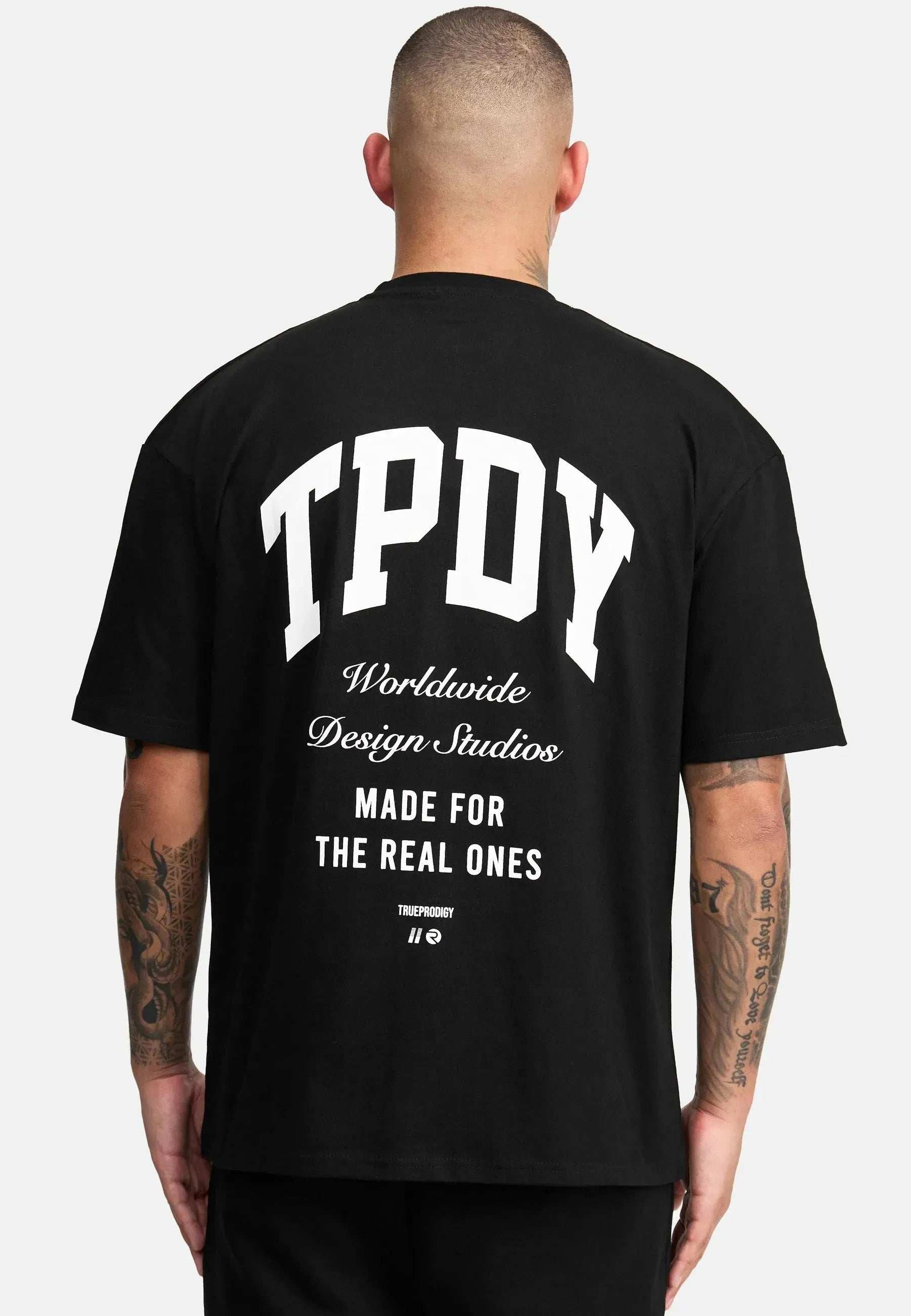 Eine Person mit rasiertem Kopf und Tattoos auf dem linken Arm trägt ein schwarzes „Elijah Oversized T-Shirt mit Druck“ von trueprodigy. Auf der Rückseite steht „TPDY Worldwide Design Studios Made For The Real Ones“, was sich mühelos mit dem schlichten weißen Hintergrund verbindet und einen kühnen Stil zeigt.