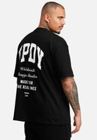 Ein Mann steht seitlich in einem Elijah Oversized T-Shirt mit Druck von trueprodigy. Auf dem fettgedruckten weißen Rücken des schwarzen T-Shirts steht in kleinerem Text und Grafiken „WPOY Worldwide Design Studio Made for the Real Ones“. Das schwere Trikot fällt gut und zeigt seine Armtattoos.