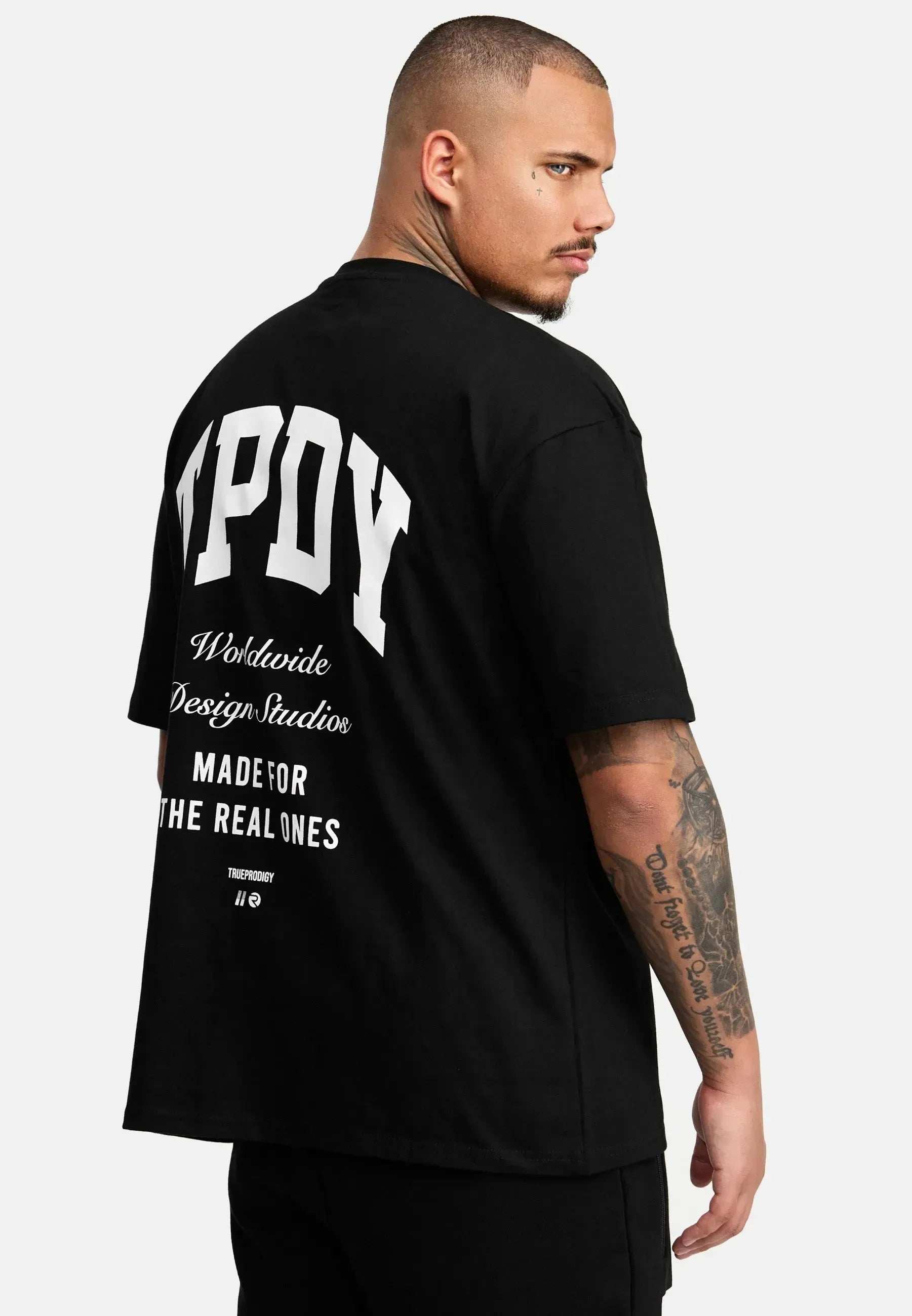 Ein Mann steht seitlich in einem Elijah Oversized T-Shirt mit Druck von trueprodigy. Auf dem fettgedruckten weißen Rücken des schwarzen T-Shirts steht in kleinerem Text und Grafiken „WPOY Worldwide Design Studio Made for the Real Ones“. Das schwere Trikot fällt gut und zeigt seine Armtattoos.