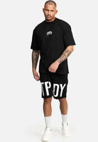 Vor einem hellgrauen Hintergrund steht ein Mann in einem übergroßen Trueprodigy Elijah T-Shirt mit Aufdruck und schwarzen Shorts und zeigt Tattoos an Armen und Beinen. Er vervollständigt den Look mit weißen Socken und Turnschuhen.