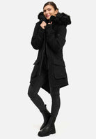 Die Person trägt den Damenparka mit Kapuze und Kunstfell von trueprodigy Harvey F, dazu dunkle Jeans und schwarze Stiefel. Sie steht vor einem schlichten Hintergrund, lächelt leicht und greift nach den Rändern der Kapuze.