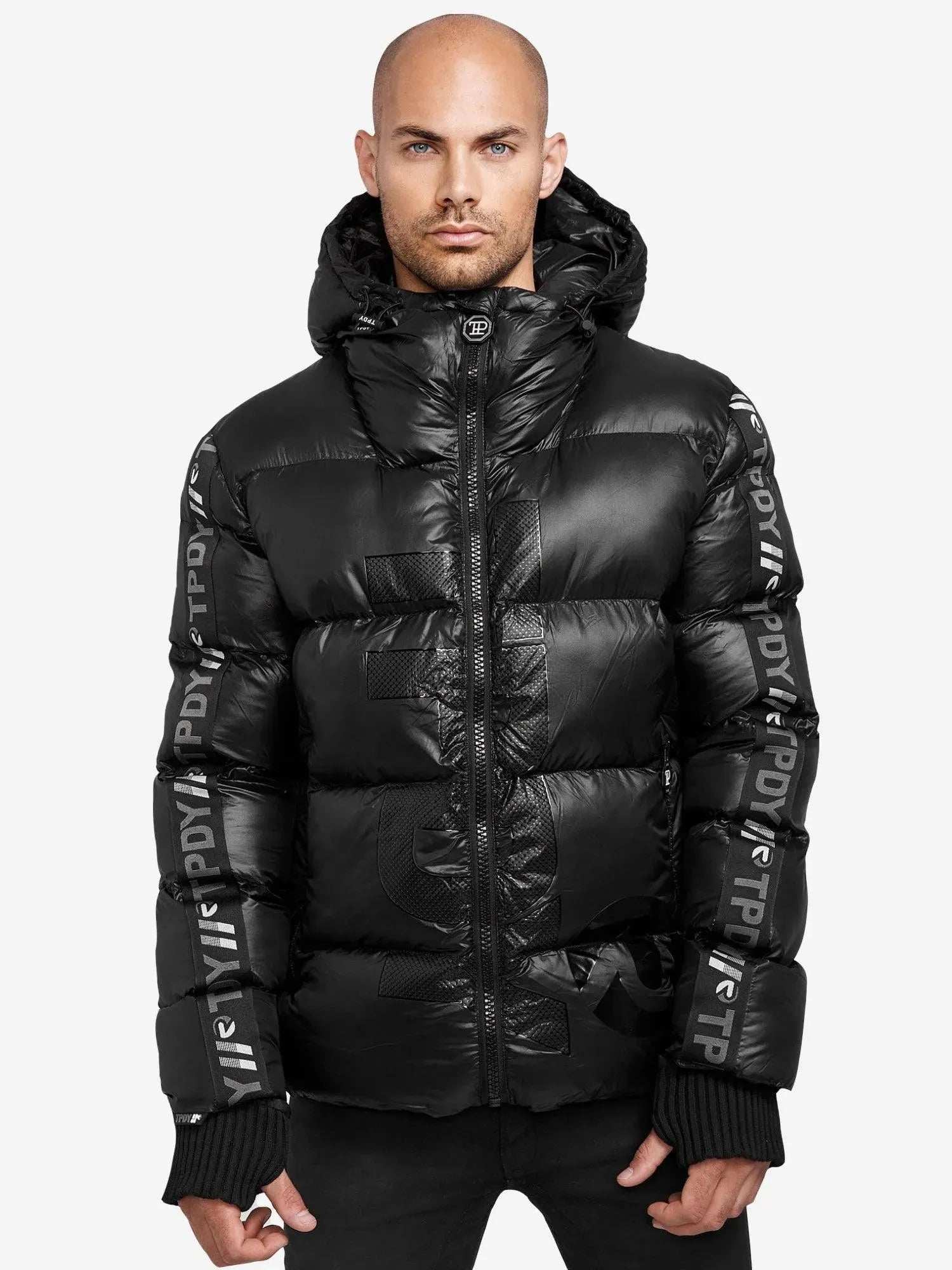 Eine kahlköpfige Person mit Gesichtsbehaarung trägt die Logan Pufferjacke mit Logo von trueprodigy. Diese glänzende schwarze Jacke mit PDYN- und PTP-Branding an Ärmeln und Brust hebt sich mit ihrer markanten Silhouette von einem schlichten weißen Hintergrund ab.