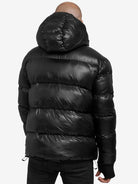 Die Person in einer glänzenden schwarzen Logan Pufferjacke mit Logo von trueprodigy, mit horizontalen Nähten und einer Kapuze, blickt von der Kamera weg. Diese stylische Jacke ergänzt dunkle Hosen vor dem schlichten weißen Hintergrund.