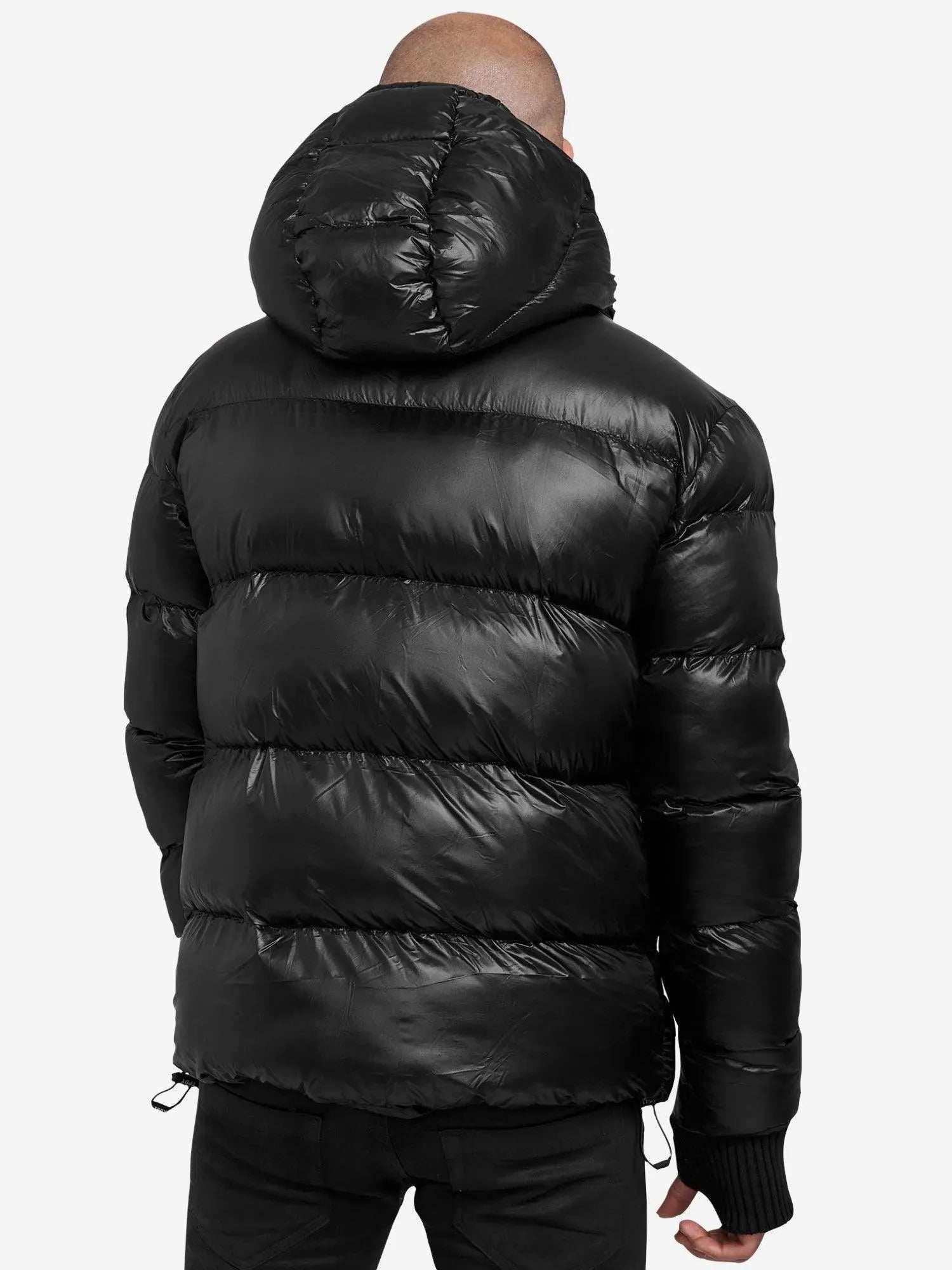 Die Person in einer glänzenden schwarzen Logan Pufferjacke mit Logo von trueprodigy, mit horizontalen Nähten und einer Kapuze, blickt von der Kamera weg. Diese stylische Jacke ergänzt dunkle Hosen vor dem schlichten weißen Hintergrund.