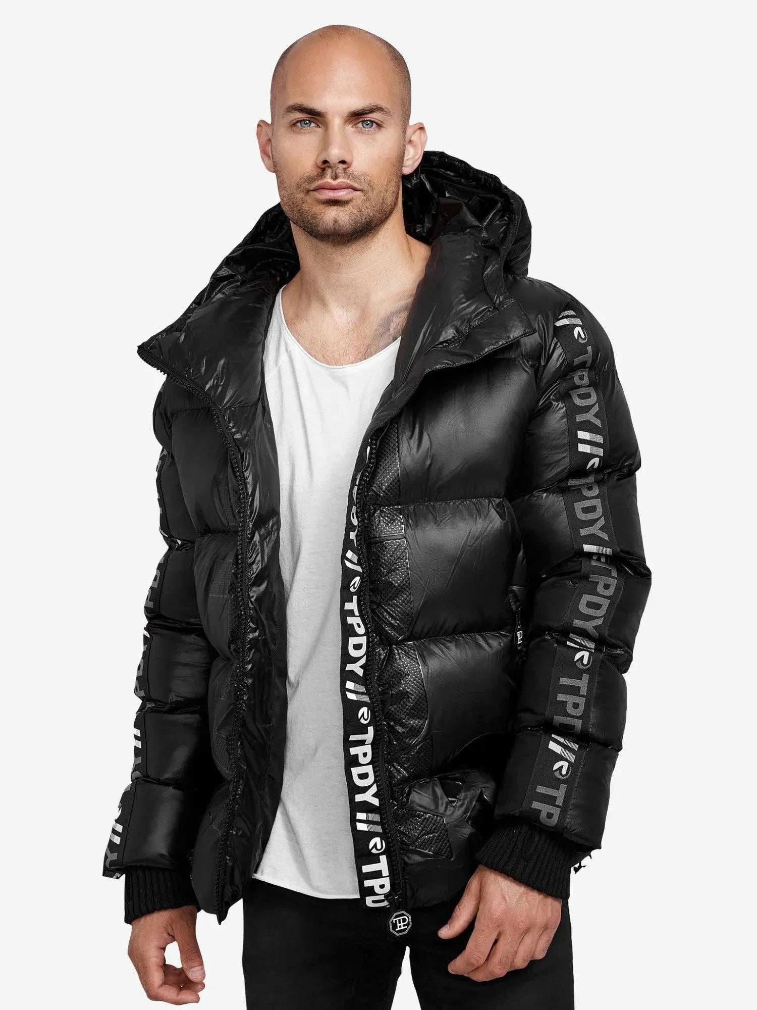 Ein kahlköpfiger Mann trägt die Logan Pufferjacke mit Logo von trueprodigy, mit aufgedrucktem Ärmeltext und Kapuze. Er hat einen leichten Bart und steht vor einem schlichten weißen Hintergrund. Unter seiner stylischen Jacke ist ein weißes T-Shirt zu sehen.