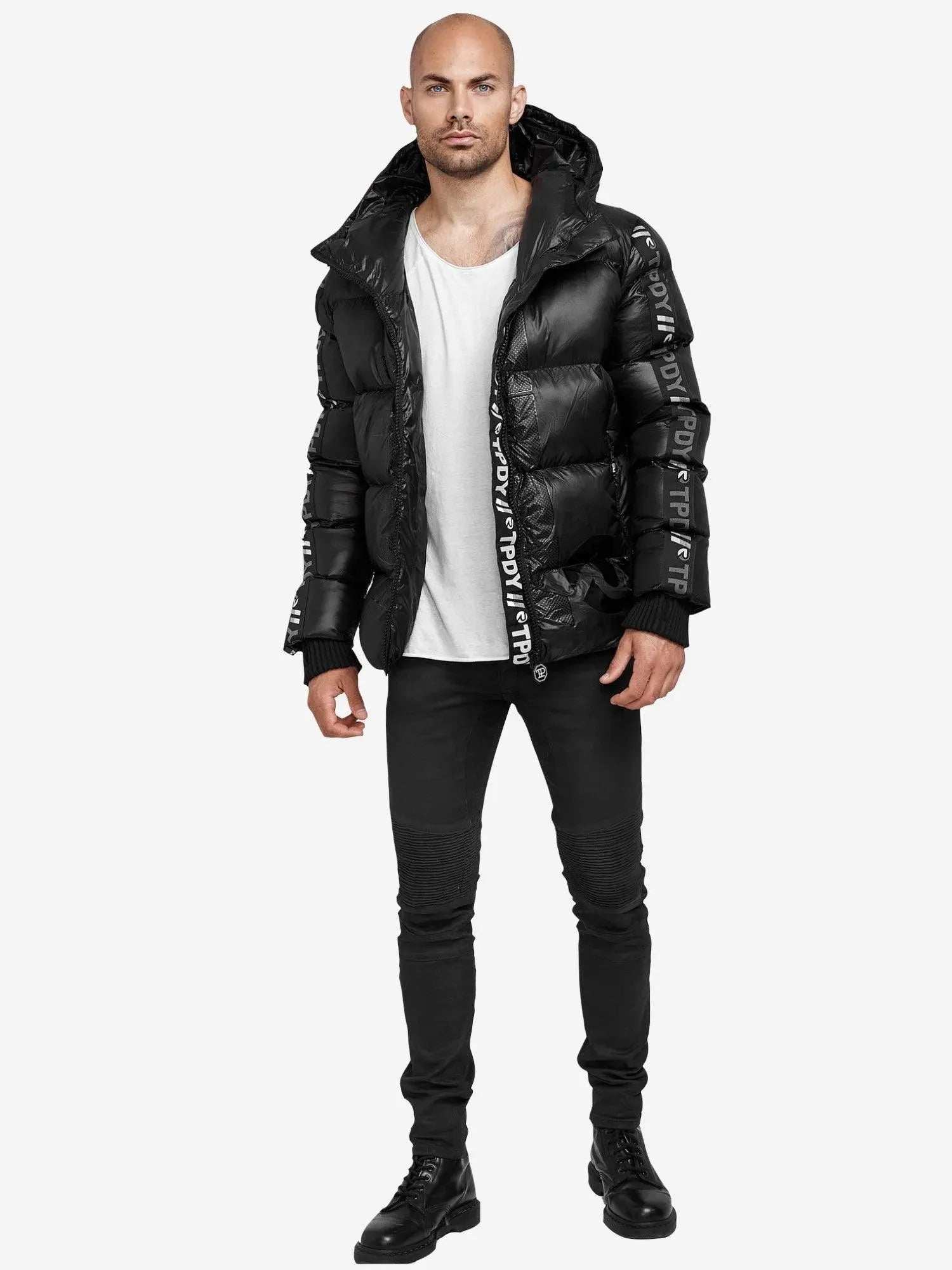 Eine Person, die eine schwarze Trueprodigy Logan Pufferjacke mit Logo trägt, steht vor einem schlichten weißen Hintergrund und trägt außerdem ein weißes T-Shirt, schwarze Jeans und schwarze Stiefel.