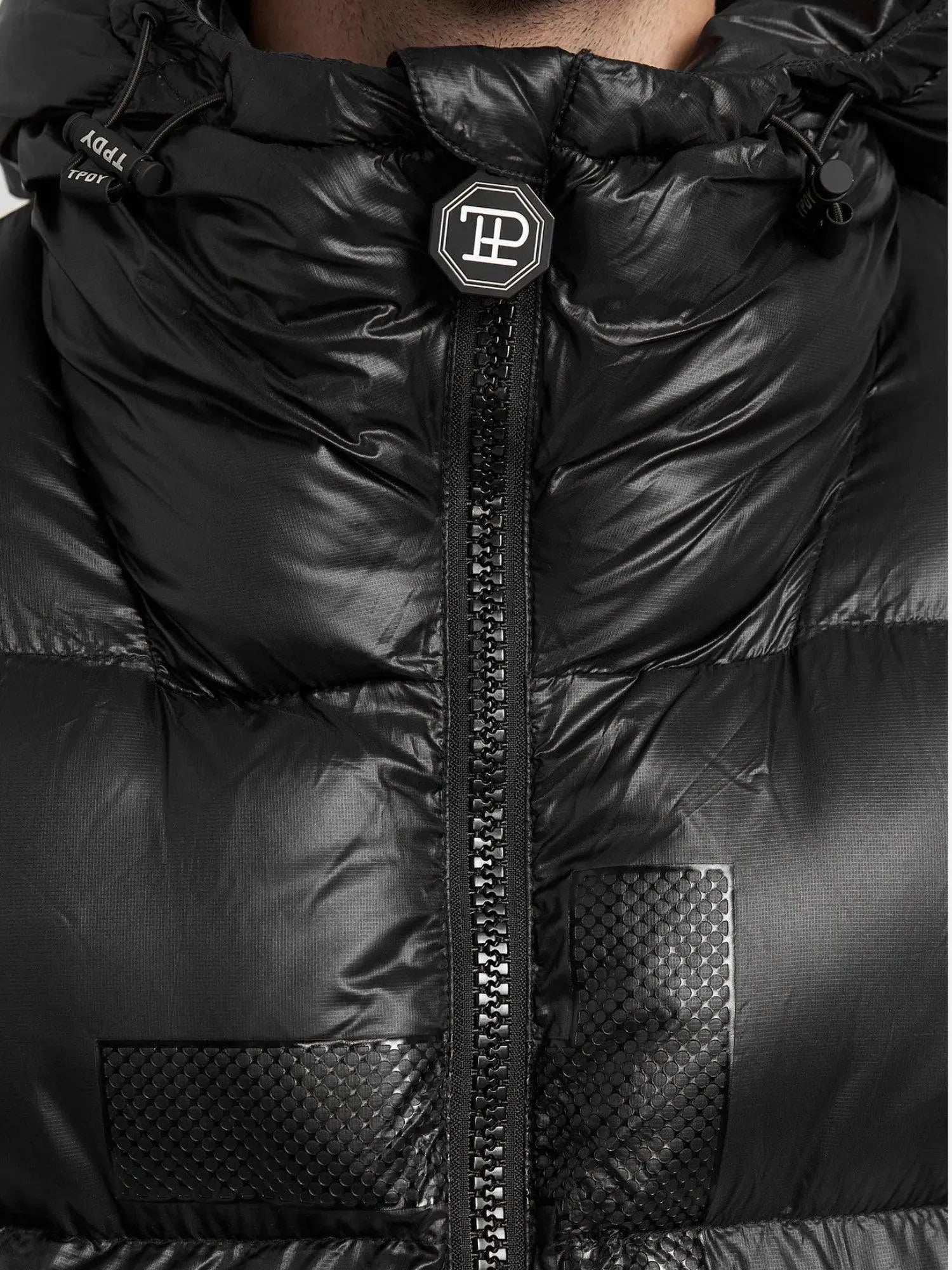 Eine Nahaufnahme einer Person, die die Trueprodigy Logan Pufferjacke mit Logo in Schwarz trägt, zeigt ihre Daunenfüllung, ihr glänzendes Finish und ihr strukturiertes Mitteldesign mit einem vertikalen Reißverschluss. Die Kapuze verfügt über Kordeln und ein rundes Logo-Abzeichen vervollständigt die schlanke Silhouette.