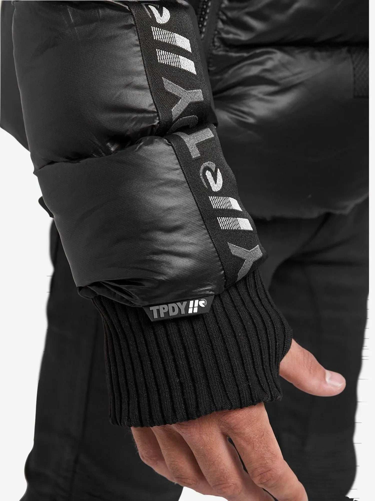 Nahaufnahme einer Person, die die Logan Pufferjacke mit Logo von trueprodigy in schlichtem Schwarz trägt, mit dem „TPDY“-Branding auf dem Ärmel und einem schwarzen Rippbündchen. Diese wattierte Jacke hat eine glänzende Textur, die ihren modernen Stil und die Logan-Silhouette unterstreicht.