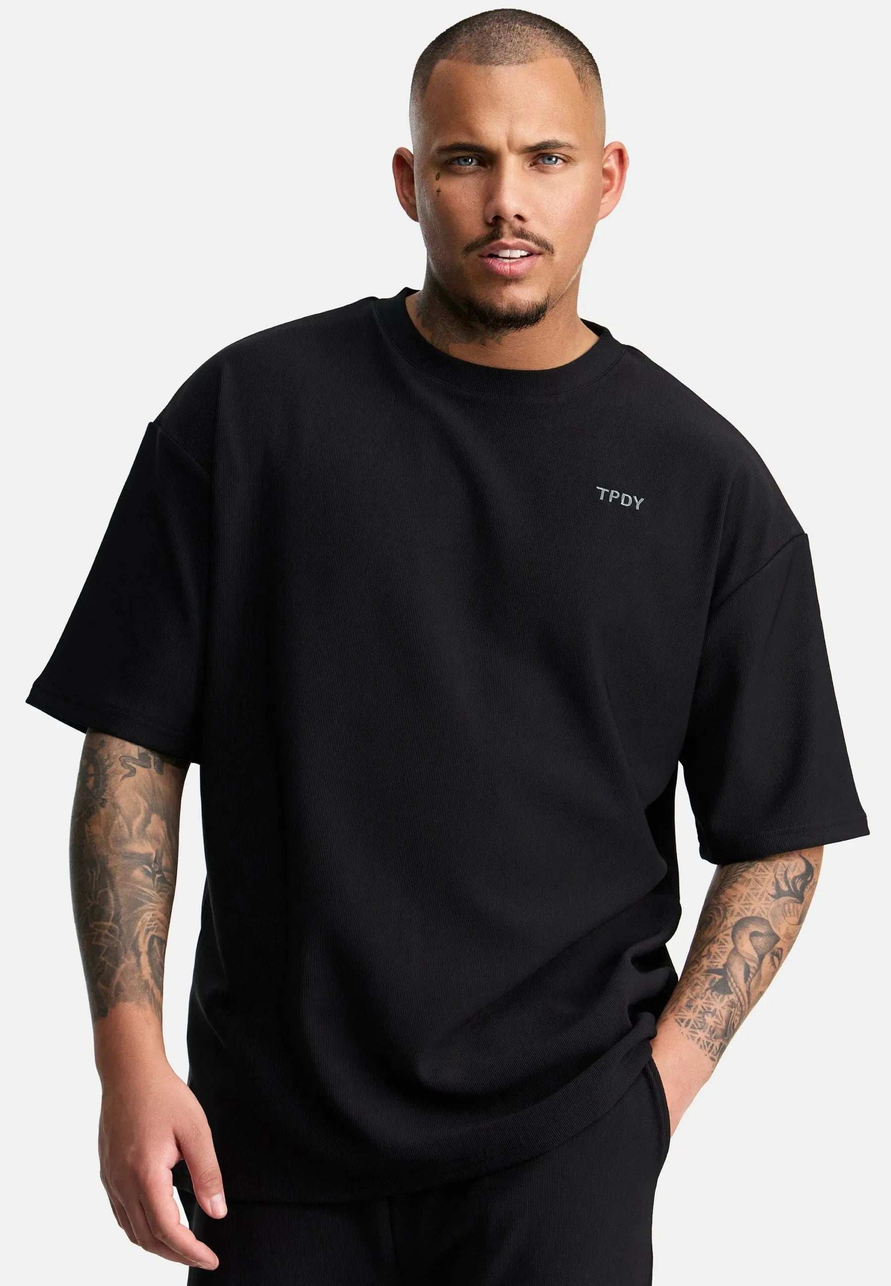 Ein Mann mit Tattoos und gestutztem Bart steht vor einem schlichten Hintergrund und trägt das Trueprodigy Mateo Oversized T-Shirt mit Stickerei mit „TPDY“ auf der Brust. Seine linke Hand steckt in der Tasche und er blickt direkt in die Kamera.