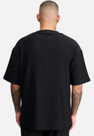 Eine Person mit rasiertem Kopf trägt von hinten gesehen das Mateo Oversized T-Shirt mit Stickerei von trueprodigy. Ihre tätowierten Arme betonen ihren Look vor einem schlichten, hellen Hintergrund.