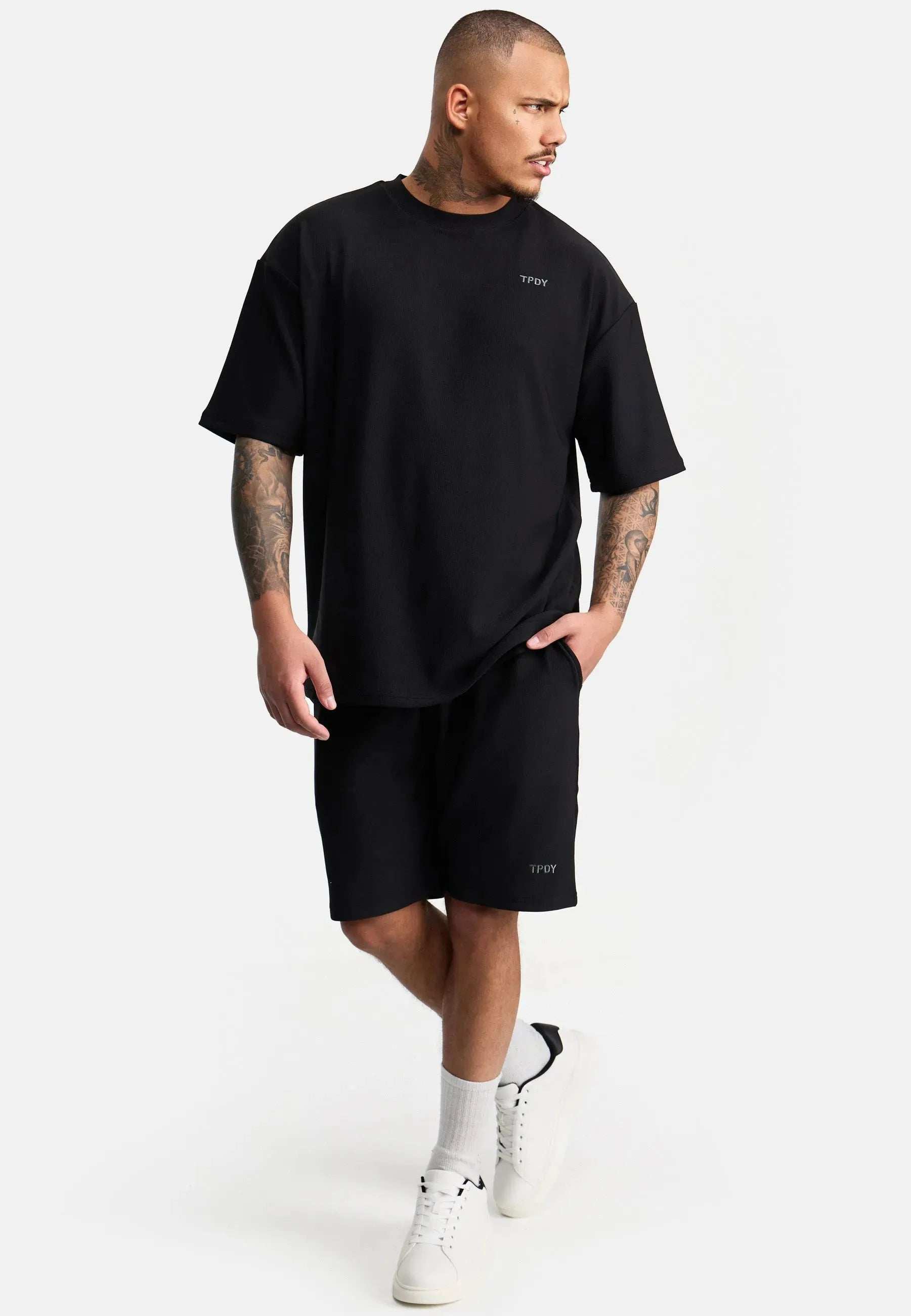 Ein Mann mit kurzen Haaren und Armtattoos trägt ein übergroßes T-Shirt mit Stickerei von trueprodigy Mateo und passende Shorts. Mit weißen Socken und Turnschuhen strahlt er vor einem schlichten weißen Hintergrund Selbstvertrauen aus und zeigt mühelosen Stil.