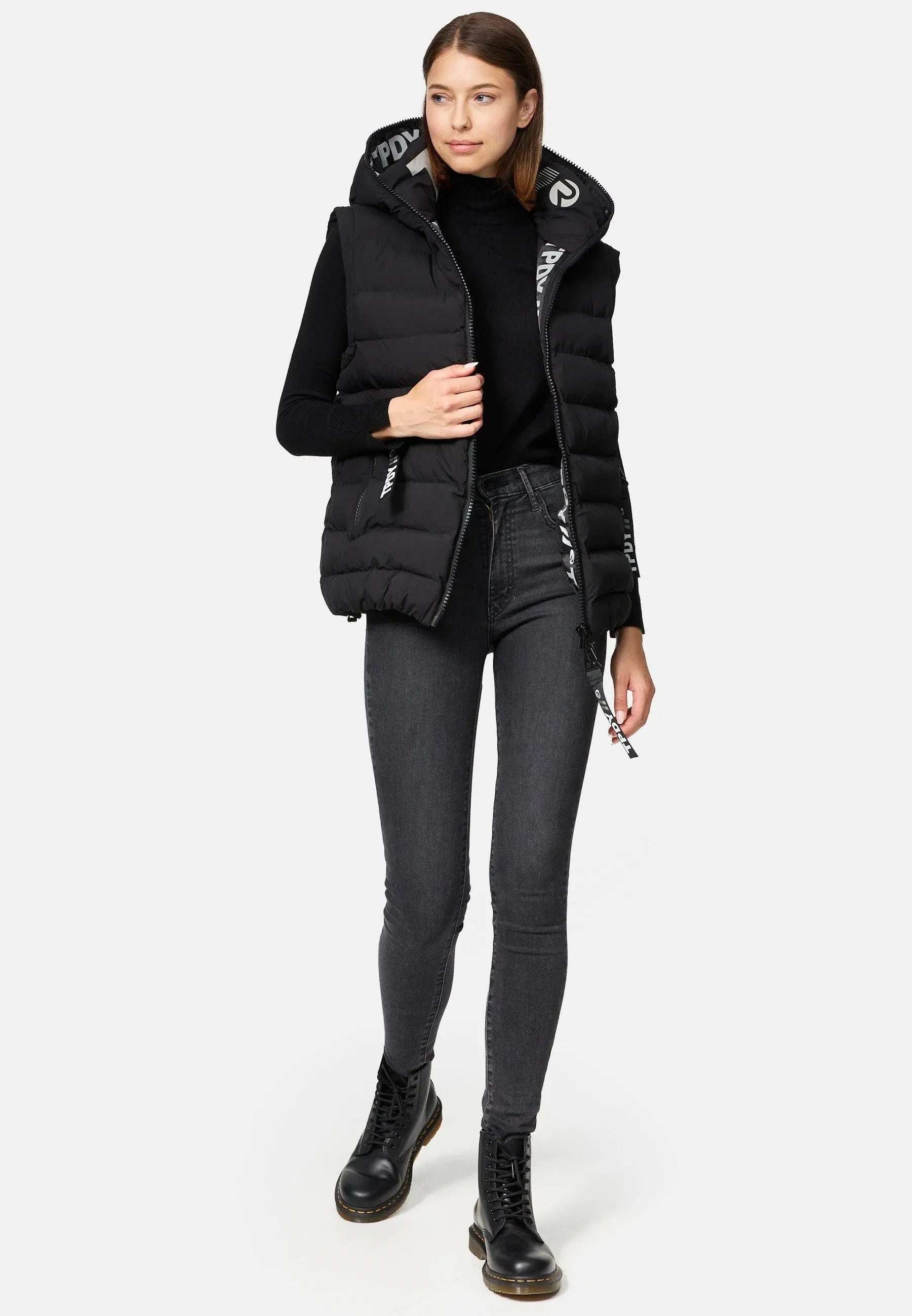 Eine Person trägt eine schwarze Damenweste mit Kapuze von Trueprodigy Matthew F über einem langärmligen schwarzen Oberteil und dunklen Jeans. Sie hat schwarze Schnürstiefel an und steht vor einem einfarbigen Hintergrund mit den Händen in der Nähe des Reißverschlusses der Weste, auf dem ein dezentes Trueprodigy-Logo zu sehen ist.