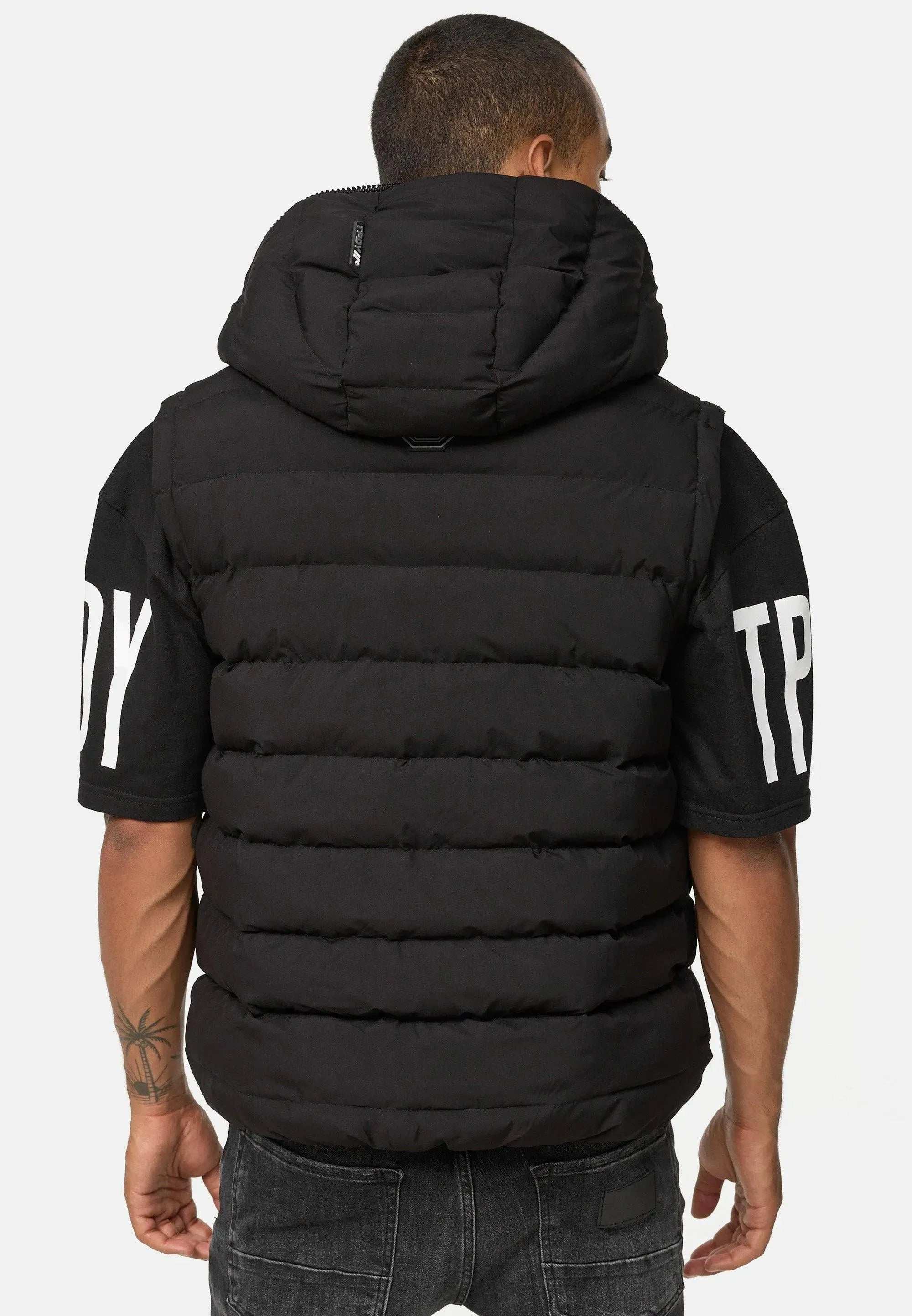 Eine Person mit kurzen Haaren blickt abgewandt und trägt eine schwarze Trueprodigy Matthew Pufferweste mit Kapuze über einem schwarzen Trueprodigy-Kurzarmshirt mit weißen Logodetails an den Ärmeln und vervollständigt den Look mit dunklen Jeans.