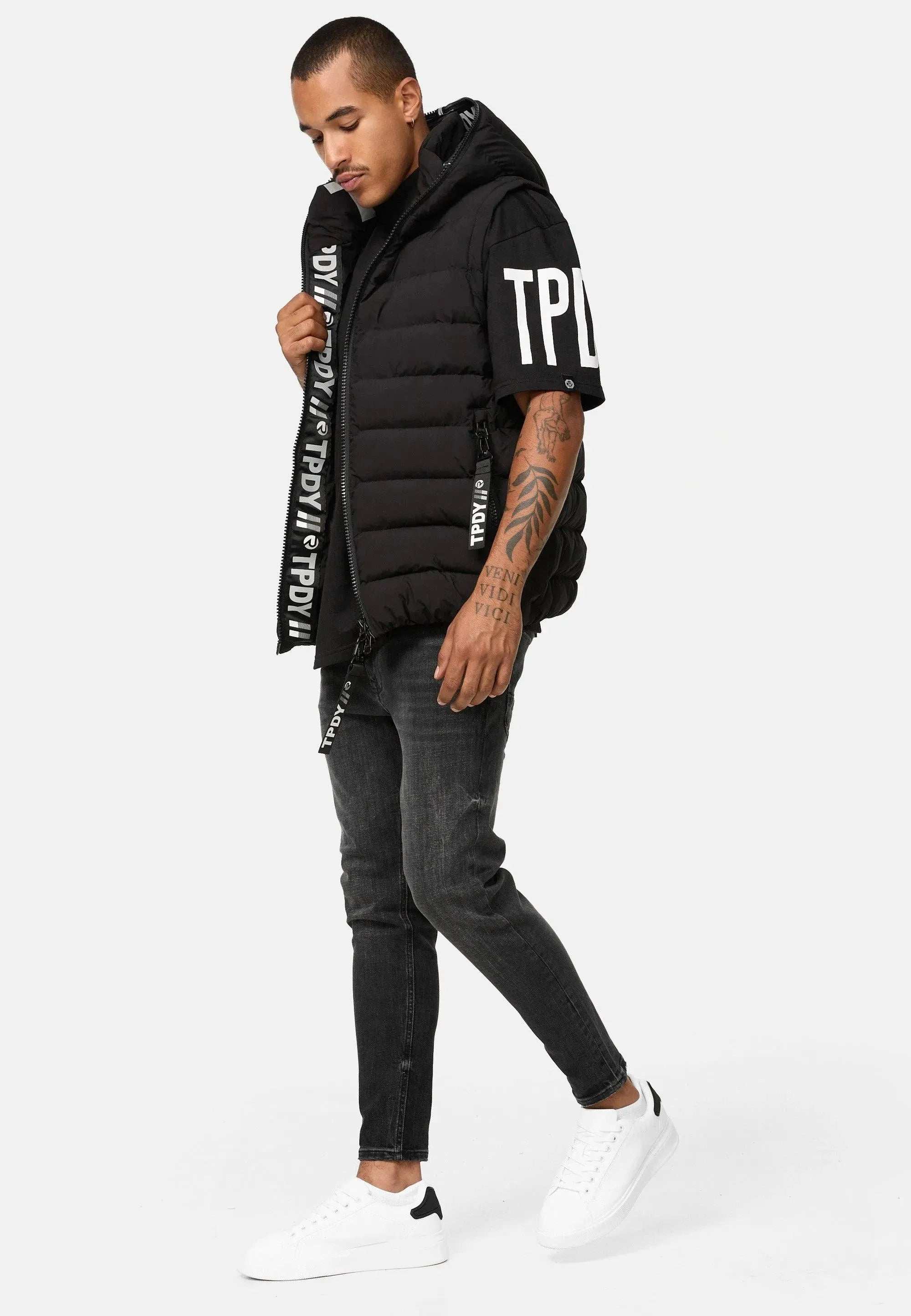 Ein Mann in einer ärmellosen, schwarzen trueprodigy Herrenweste mit Logodetails steht mit der linken Hand in der Tasche. Er trägt außerdem ein schwarzes trueprodigy T-Shirt, dunkle Jeans und weiße Turnschuhe. Auf seinem linken Arm sind Tattoos zu sehen.