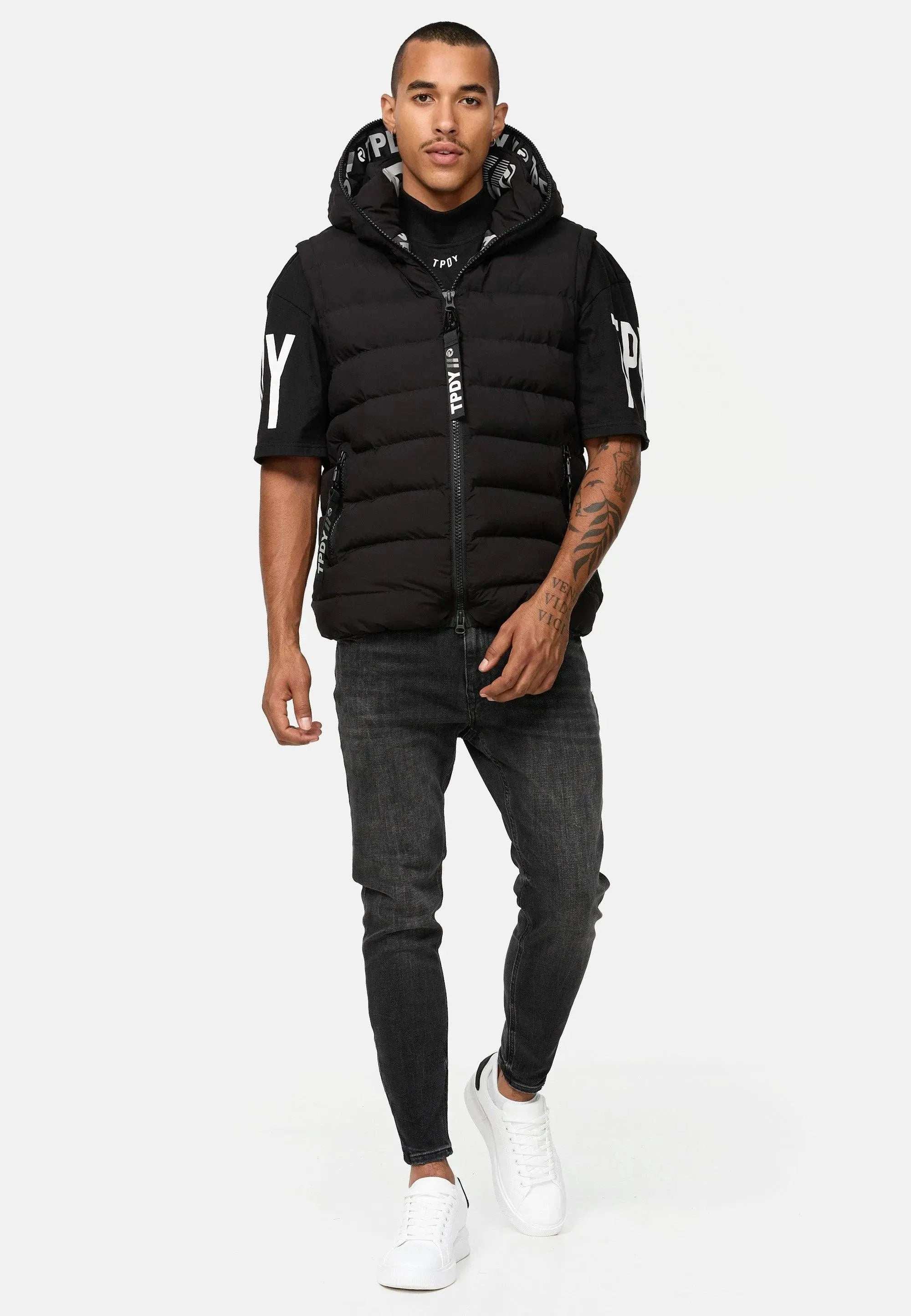 Eine Person trägt selbstbewusst eine schwarze Trueprodigy Matthew Pufferweste mit Kapuze, kombiniert mit einem schwarzen T-Shirt mit Logodetails an den Ärmeln, schwarzen Jeans und weißen Sneakers vor einem schlichten weißen Hintergrund.