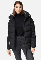Eine Frau trägt die Murphy F Matte Steppjacke mit Kapuze von trueprodigy mit Reißverschluss vorne und wasserabweisendem Design und steht vor einem schlichten weißen Hintergrund. Ihr braunes Haar fällt über die schwarze Steppjacke, die sie über einem Rollkragenpullover und dunklen Jeans trägt.