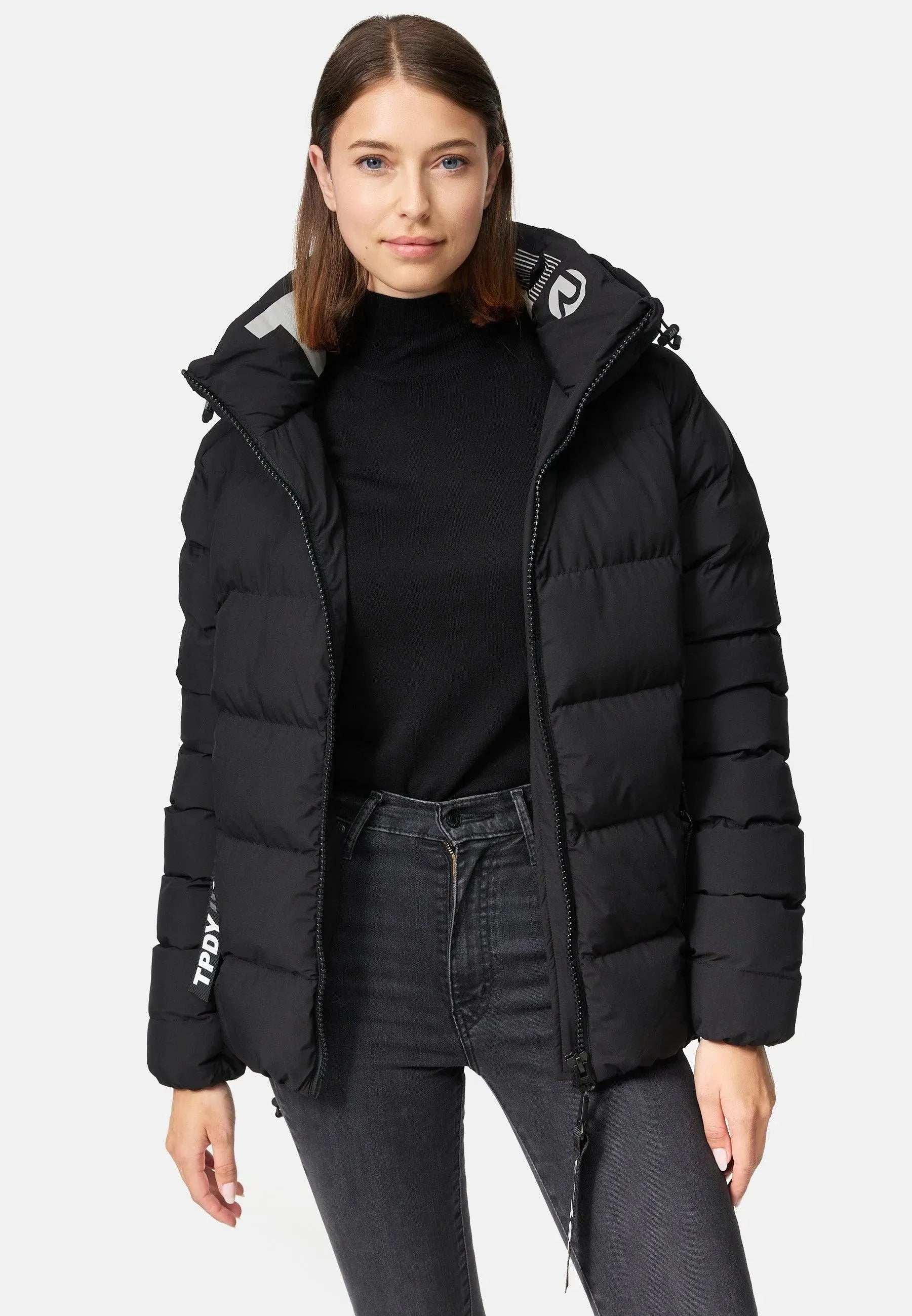 Eine Frau trägt die Murphy F Matte Steppjacke mit Kapuze von trueprodigy mit Reißverschluss vorne und wasserabweisendem Design und steht vor einem schlichten weißen Hintergrund. Ihr braunes Haar fällt über die schwarze Steppjacke, die sie über einem Rollkragenpullover und dunklen Jeans trägt.