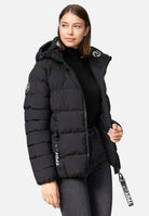 Eine Person trägt eine schwarze Steppjacke mit Kapuze von trueprodigy Murphy F Matte, mit „TPDY“ auf den Reißverschlusslaschen und einem Logo auf dem Ärmel, dazu einen schwarzen Rollkragenpullover und Jeans. Die wasserabweisende Jacke hat eine feste Kapuze. Sie blickt zur Seite.