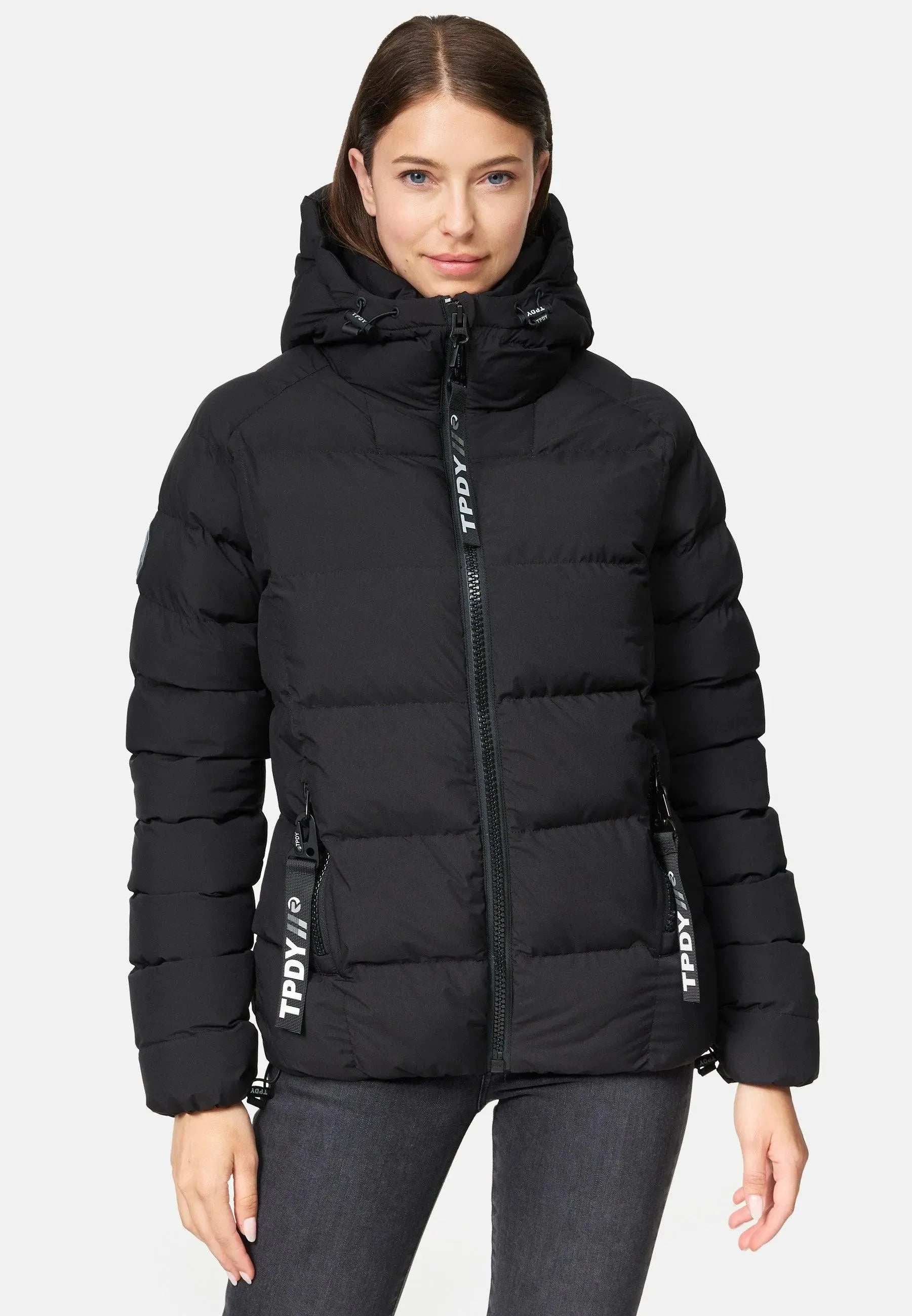 Eine Person in einer schwarzen Murphy F Matte Damenjacke von trueprodigy mit fester Kapuze steht vor einem schlichten hellen Hintergrund. Die wasserabweisende Jacke hat Reißverschlusstaschen mit der Aufschrift „TPDY“. Sie blickt geradeaus, mit neutralem Gesichtsausdruck.