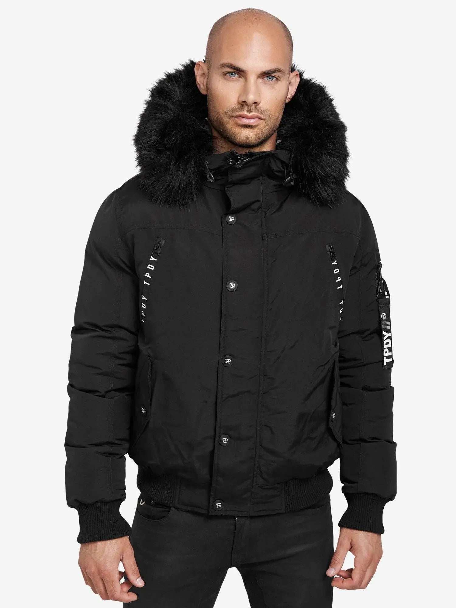 Eine Person mit rasiertem Kopf trägt den Noah Winterparka mit Kapuze und Kunstfell von trueprodigy, eine schwarze Pufferjacke mit einem großen Kunstfellbesatz auf der Kapuze. Sie ähnelt einer Bomberjacke mit Druckknöpfen und Textstreifen und hebt sich vom schlichten weißen Hintergrund ab.