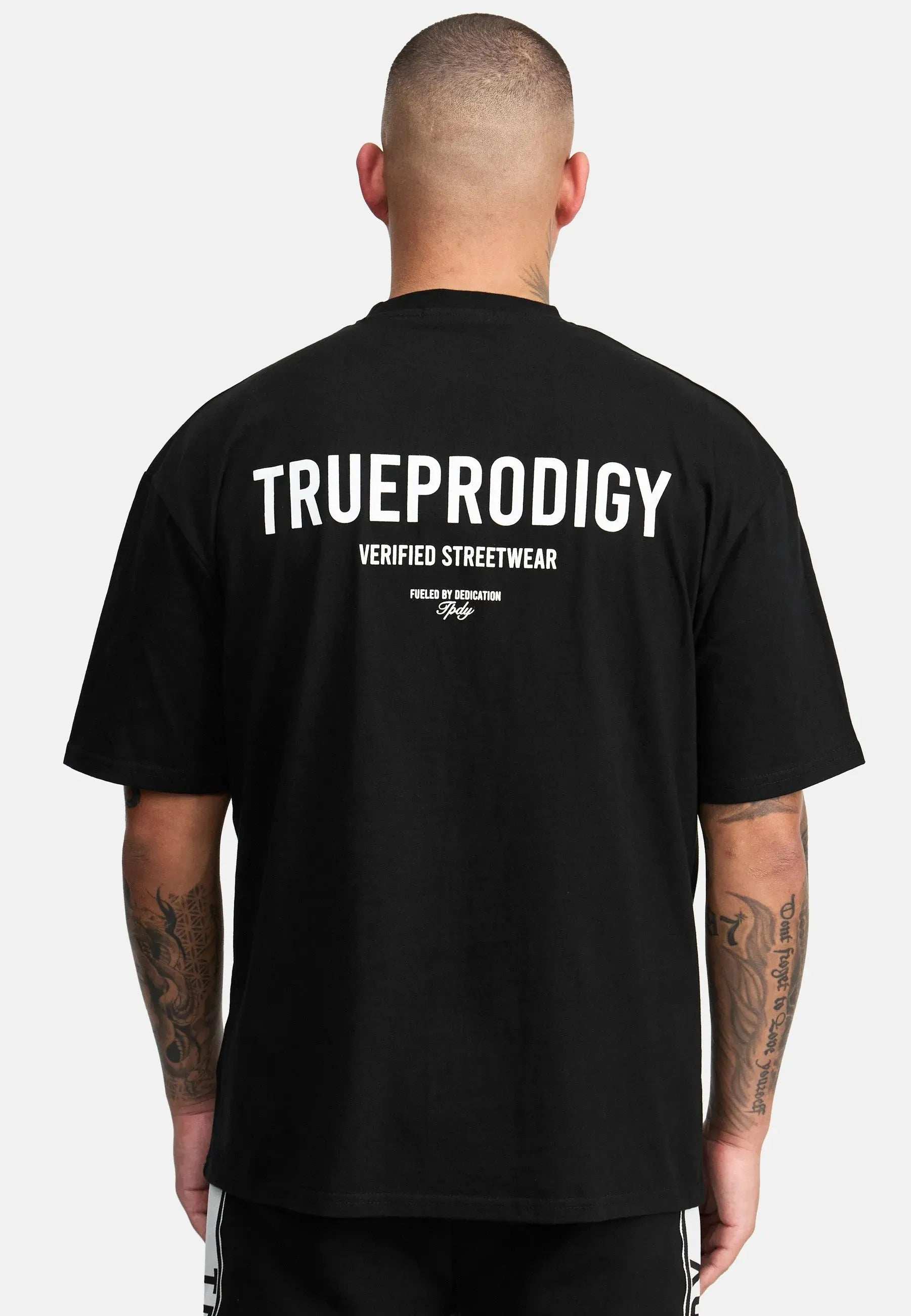 Eine Person mit kurzen Haaren blickt abgewandt und trägt ein schwarzes TrueProdigy Oliver Oversized T-Shirt aus schwerem Jersey. Auf der Rückseite ist in Weiß „TRUEPRODIGY VERIFIED STREETWEAR“ aufgedruckt. Ihre tätowierten Arme heben sich vor einem schlichten weißen Hintergrund ab.