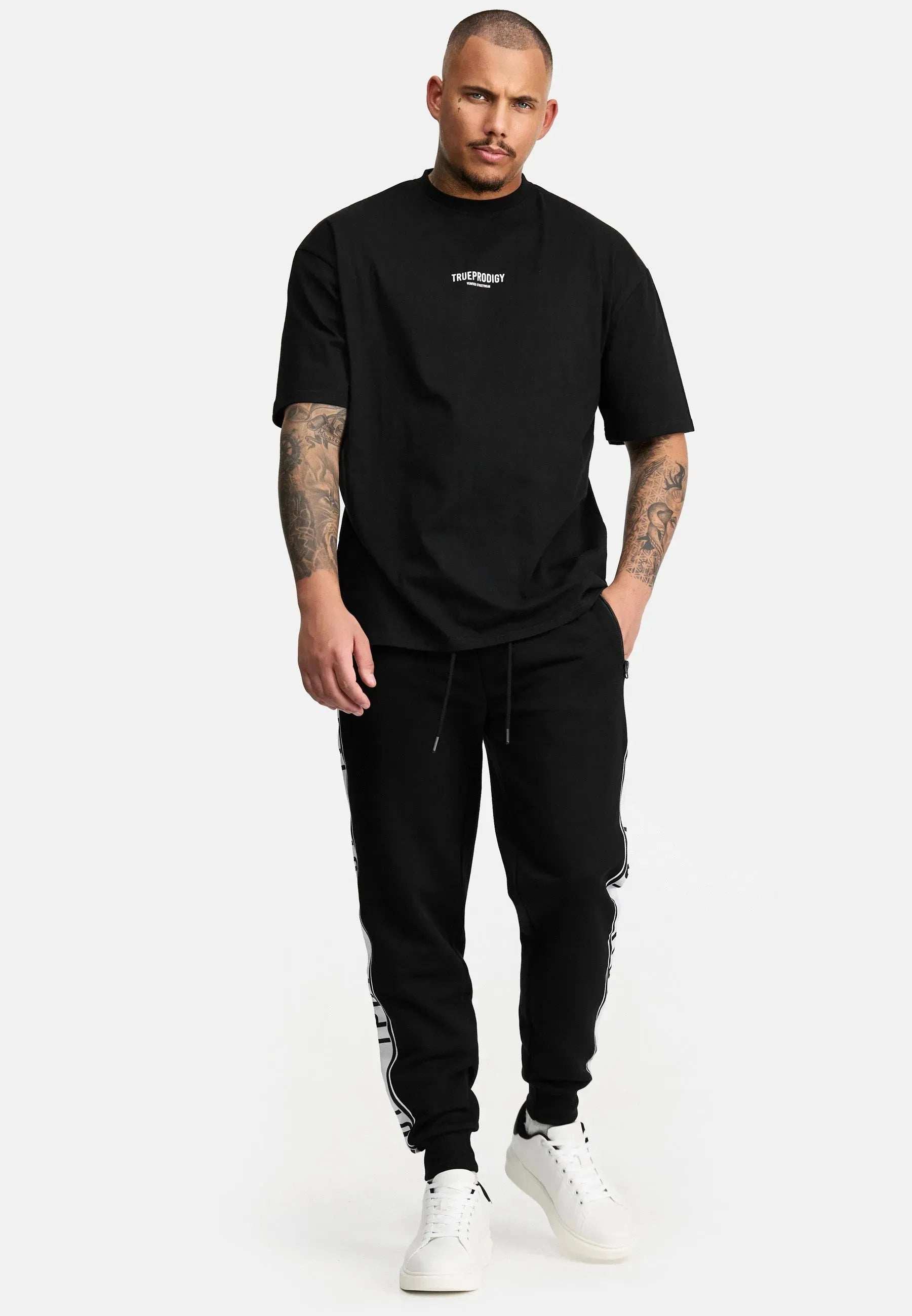 Ein Mann mit kurzen Haaren steht vor einem weißen Hintergrund und trägt ein schwarzes Oliver Oversized T-Shirt mit Druck von trueprodigy, dazu schwarze Jogginghosen und weiße Turnschuhe. Tätowierte Arme ruhen an seinen Seiten, während er mit neutralem Gesichtsausdruck nach vorne blickt.