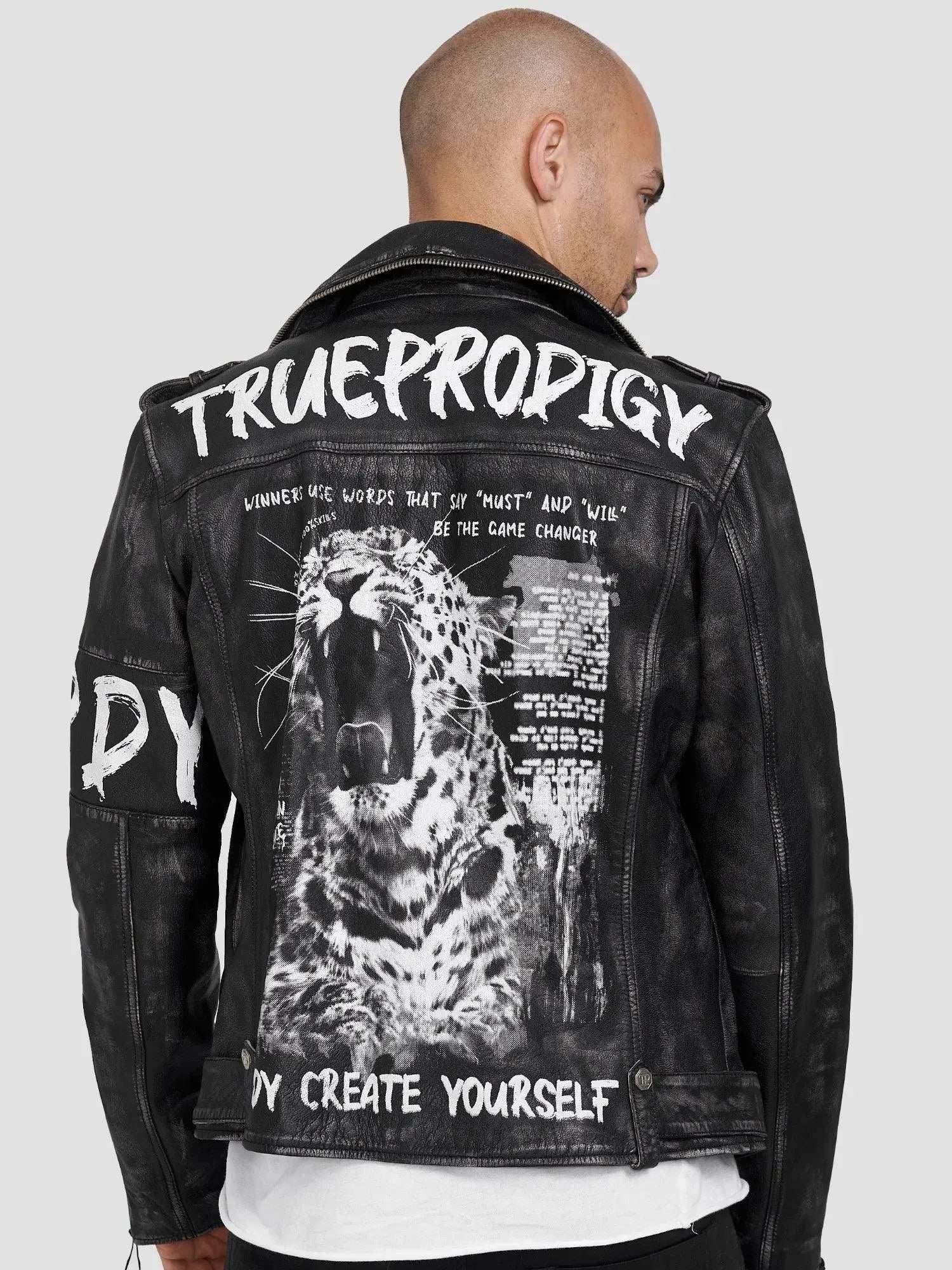 Ein kahlköpfiger Mann trägt die Lederjacke „Russell“ von TrueProdigy im Biker-Look mit weißem Text und grafischem Aufdruck eines brüllenden Leoparden auf dem Rücken. Sie hat ein Distressed-Design auf einem schlichten grauen Hintergrund mit auffälligem Aufdruck „TRUEPRODIGY“.