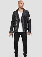 Ein Mann in der Russell-Lederjacke von trueprodigy mit dem Aufdruck „TOY CREATES YOU“ und Nieten-Akzenten steht vor einem grauen Hintergrund. Er verkörpert den Biker-Look, indem er sie mit einem weißen T-Shirt und einer schwarzen Hose kombiniert.