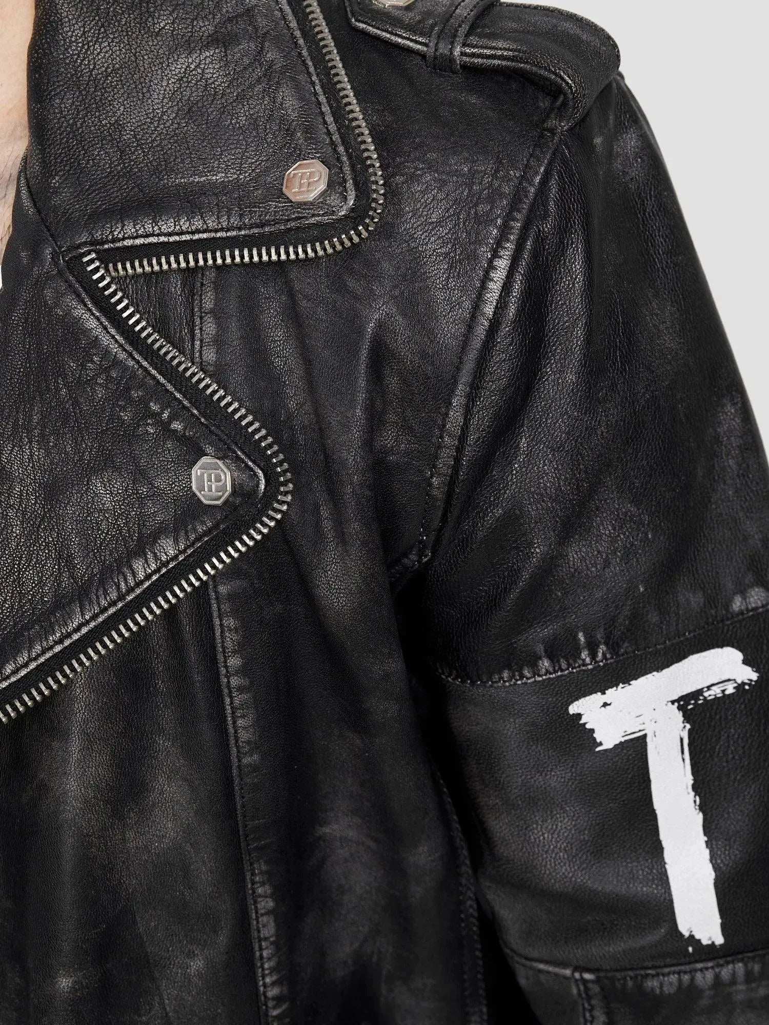 Nahaufnahme der Russell Lederjacke mit Print von trueprodigy mit silbernen Reißverschlüssen, Metallknöpfen und klassischem Biker-Look. Auf den Ärmel ist ein weißes „T“ mit sichtbarem Patchwork und Struktur aufgemalt, und eine Schulterklappe vervollständigt das kantige Design.