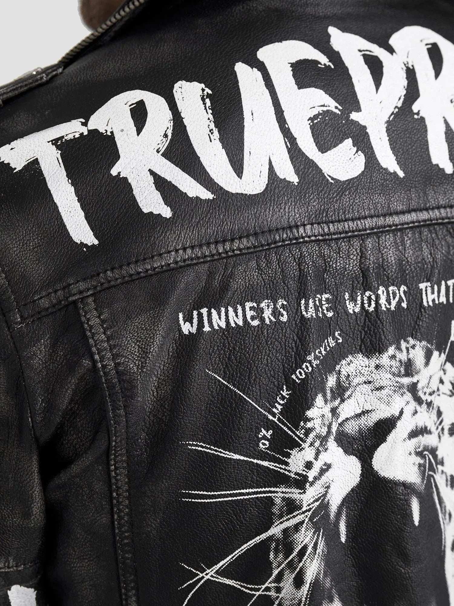 Nahaufnahme der Russell Lederjacke mit Print von trueprodigy, mit weiß gemaltem Text auf schwarzem Leder. Oben steht „TRUEPR“, darunter „WINNERS USE WORDS THAT“, mit einer Grafik eines brüllenden Löwen, überlagert mit „DO IT LIKE 100% SKILLS“. Fügen Sie noch einen kantigen Biker-Look hinzu.